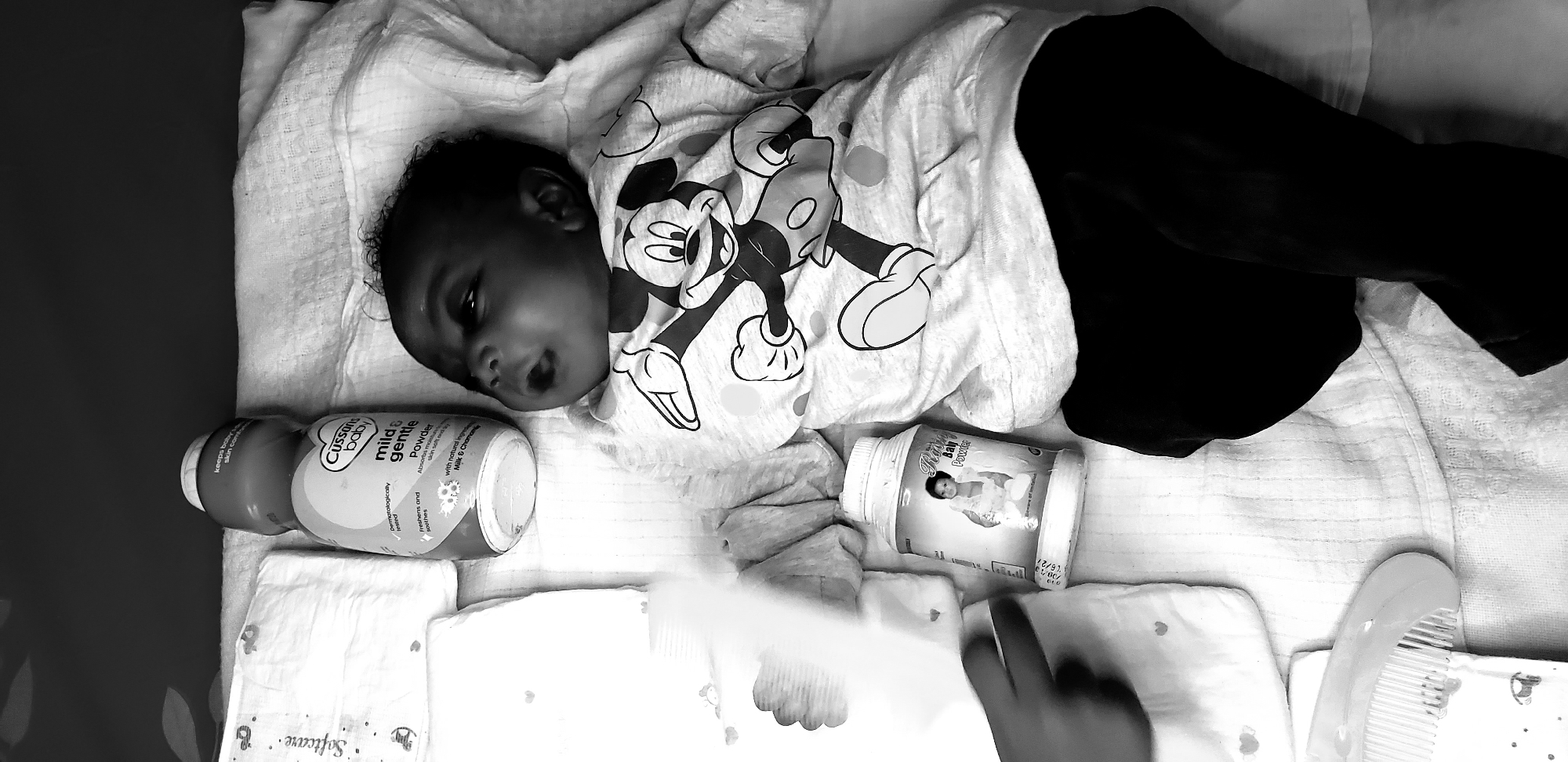 A baby in a Mickey Mouse shirt lies on a white cloth, surrounded by baby products like Cussons Baby Mild & Gentle Powder and Ram Day Power. A package labeled "Softcare" is visible. The scene appears to be a changing area, possibly in a home in Narayi, Nigeria. A comb and diapers are present. A hand holding a baby wipe is present in the foreground.
