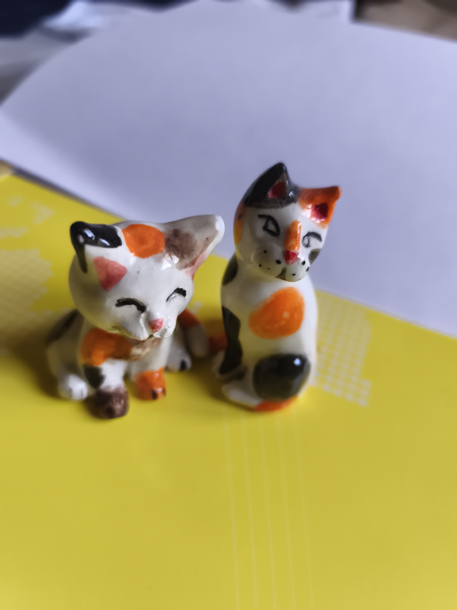 Two small, ceramic cat figurines are placed on a bright yellow surface. The figurine on the left is sitting with its eyes closed and appears to have white, orange, and black markings. The figurine on the right is also sitting and has similar markings of white, orange, and black, with its eyes open and head tilted. The background is out of focus, showing a white surface. There is no discernible location in the city of Saint-Denis, France.