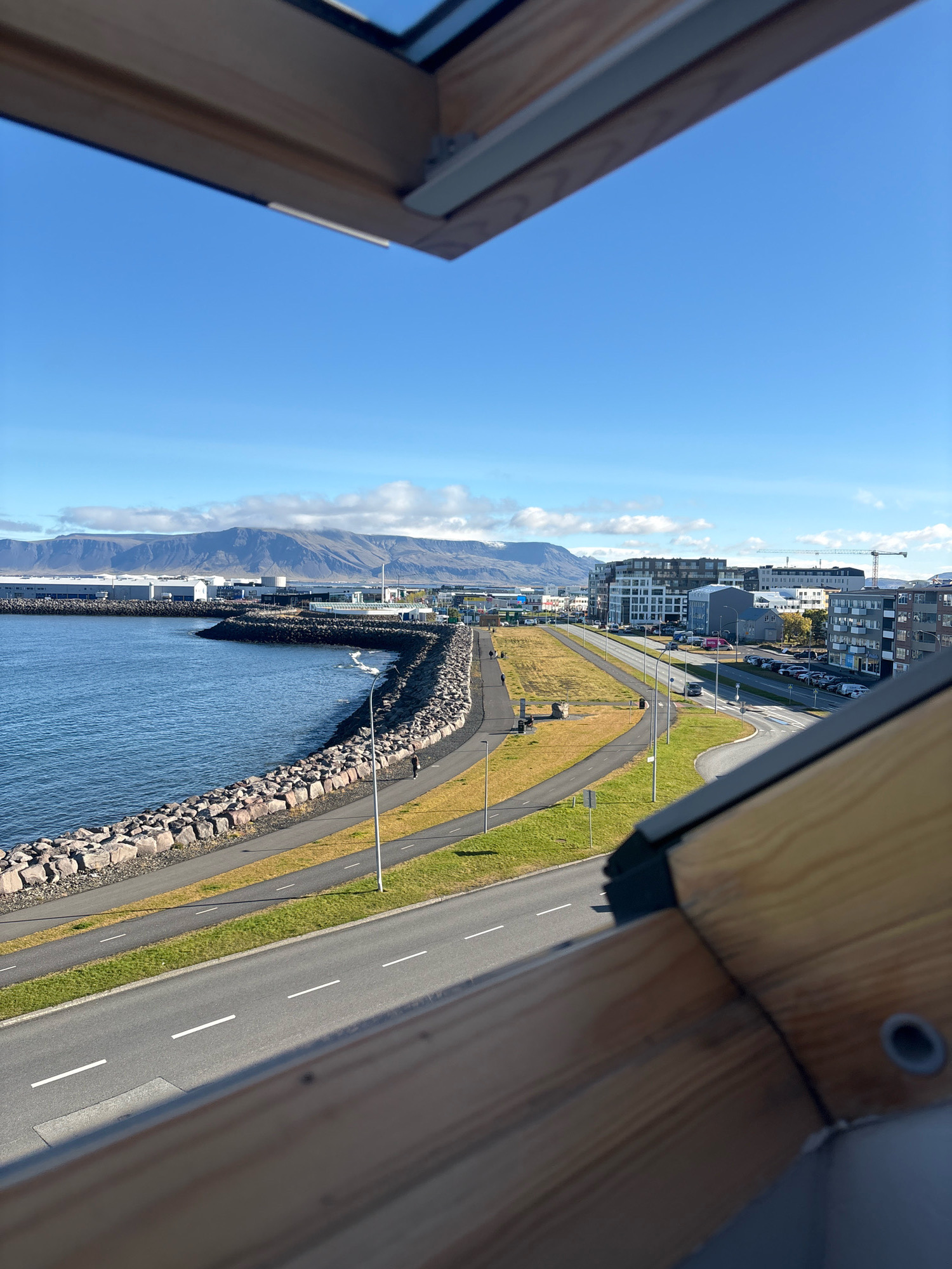 This is a high-angle, slightly blurred photographic view of a coastal area in Reykjavik, Iceland, likely from an elevated vantage point such as a building or a skylight. The foreground is dominated by the wooden framing of what appears to be a window or roof structure, casting a shadow over part of the scene.
The middle ground shows a paved road with white lane markings running parallel to a rocky breakwater. Beyond the breakwater is a body of water, appearing calm and blue. A paved walkway or bike path runs alongside the breakwater, with a lone figure walking on it. Further inland, there's a strip of grass and then another paved road lined with parked cars and streetlights.
The background reveals the cityscape of Reykjavik, characterized by modern buildings of varying heights, including apartment blocks and industrial structures. A construction crane is visible in the distance. Towering over the city and extending to the horizon are majestic mountains, some of which are capped with clouds. The sky is a clear, bright blue, with a few scattered white clouds.
The overall scene depicts a harmonious blend of urban development, natural coastal landscape, and mountainous scenery, characteristic of Reykjavik. The time of day appears to be daytime, given the lighting and the clear sky.