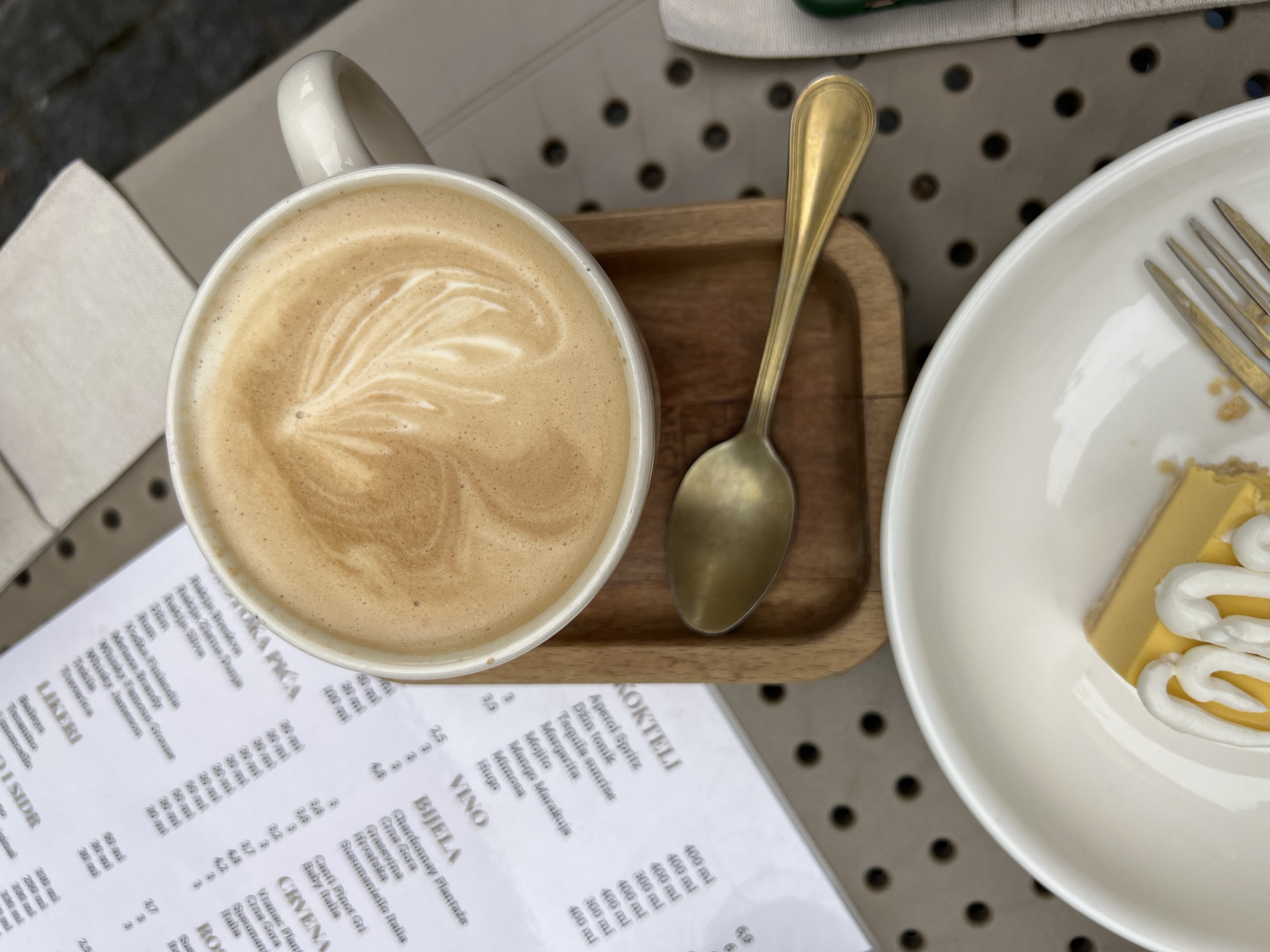 The image shows a top-down view of a table setting with a cup of latte, a small wooden tray with a golden spoon, a plate with a partially eaten dessert (possibly lemon cheesecake), and a menu card. The latte has latte art on top. The dessert is topped with swirls of whipped cream. The menu lists various drinks, including cocktails ("KOKTELI"), wines ("VINO"), liqueurs ("LIKERI"), and possibly other beverages. The menu appears to be in the Montenegrin language. A fork is on the dessert plate. The table has a light-colored, possibly perforated surface. The scene is likely outdoors, based on the natural lighting and the table surface, suggesting a cafe or restaurant setting in Podgorica, Montenegro.