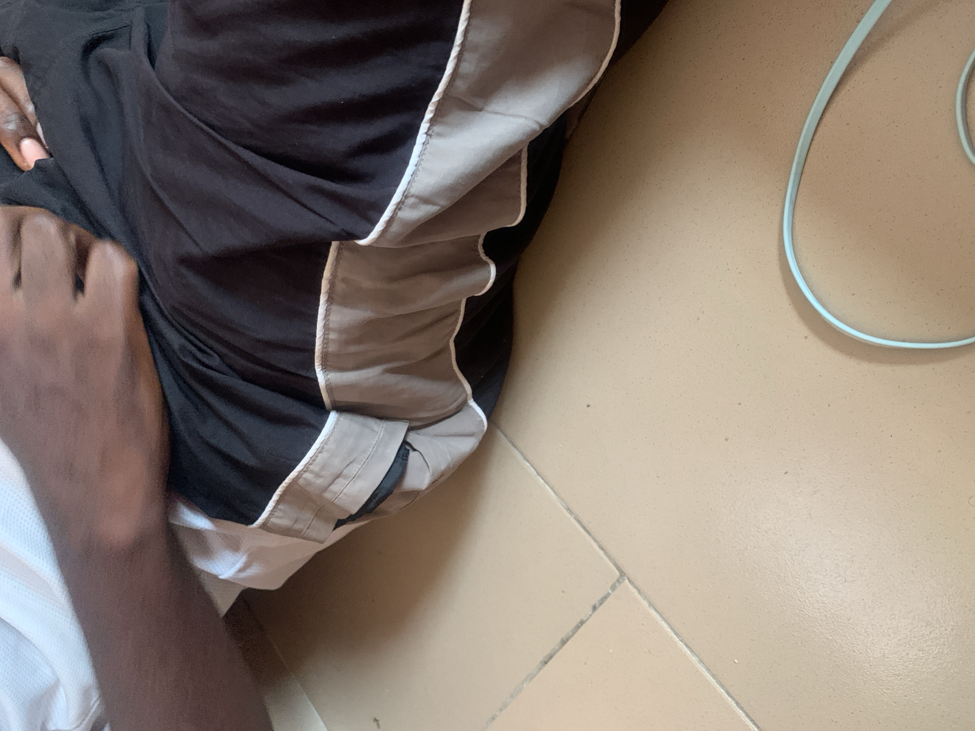 In the city of Jauro Yinu, Nigeria, this image captures a close-up of a person's lap covered by black and gray striped shorts, with a white shirt visible at the top. The person's arm rests on their lap. The scene is set on a tiled floor. A light blue cable is visible in the foreground.