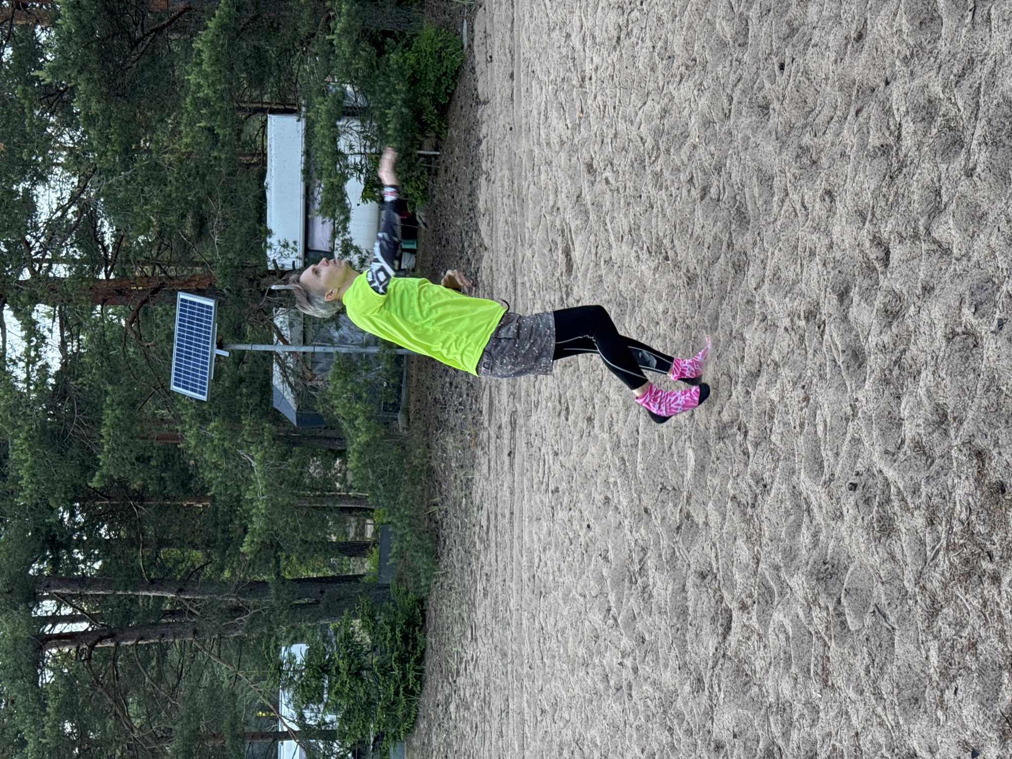 The image captures a person with short, blond hair standing on a sandy beach in Zelenaya Roshcha, Russia, in what appears to be a wooded area adjacent to a body of water. The person is wearing a bright neon green long-sleeved shirt, a patterned skirt, black leggings, and distinctive bright pink socks with a white pattern. They are in the process of throwing something, with their arm extended and body in mid-motion. In the background, there are tall pine trees, some structures that could be buildings or RVs, and a solar panel mounted on a pole. The ground is covered in coarse sand with sparse vegetation. The lighting suggests it is daytime, possibly overcast or late afternoon.