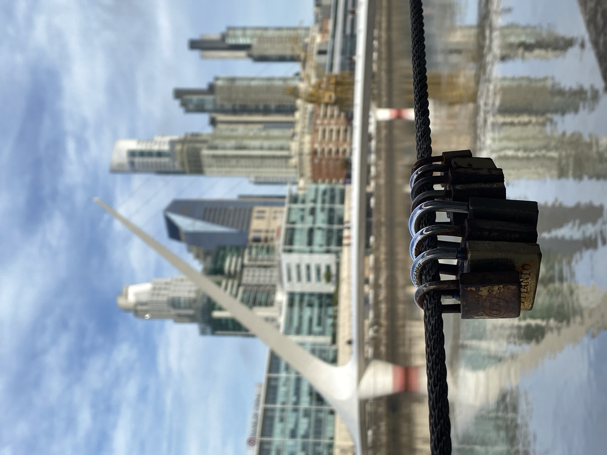 The image shows a close-up view of several padlocks attached to a black rope or cable. In the background, out of focus, is a cityscape, including the Puente de la Mujer (Woman's Bridge), a distinctive pedestrian bridge with a single mast, and modern buildings. The scene is located in Puerto Madero, Buenos Aires, Argentina, a revitalized docklands area. The padlocks appear to be love locks, often attached to bridges or other public structures as a symbol of romantic commitment. The water reflecting the buildings and sky is visible behind the locks and the rope.