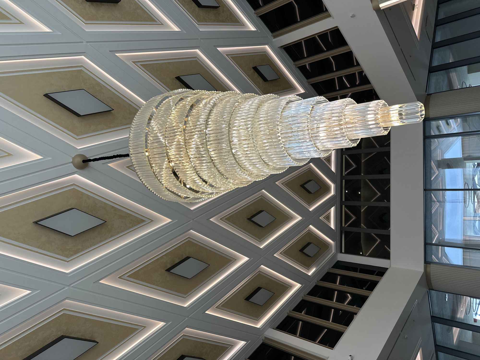 This is an eye-level, slightly upward-angled interior shot of a grand ceiling featuring a large, ornate chandelier, likely within a modern building in Kazanlak, Bulgaria.
The primary subject is a massive, multi-tiered chandelier made of numerous clear, cylindrical or rectangular glass/crystal segments arranged in rings. The chandelier tapers from a wider base to a narrower top, emanating a warm, bright golden light that reflects off its surfaces. It is suspended from a circular base, with a dark, braided cable connecting it to the ceiling.
The ceiling itself is a significant feature, adorned with a geometric pattern of large, recessed diamond-shaped panels. These panels are framed by thick, white architectural molding, within which strips of warm LED lighting are embedded, illuminating the edges of the diamond sections. The inner part of each diamond panel is a textured, light beige or gold color, and many contain a smaller, darker square fixture with a light grey or white center, possibly a speaker or an additional light source, set within a raised frame.
To the right side of the image, large windows or glass panels extend vertically, suggesting a multi-story space or an atrium. Reflections of the chandelier and the decorative ceiling are visible in these windows, along with glimpses of a blue sky and distant structures, indicating it is likely daytime. The upper right corner also shows dark, possibly reflective ceiling sections with a grid pattern, adding to the contemporary and luxurious aesthetic of the space.
No people or activities are visible. There is no discernible text in the image.