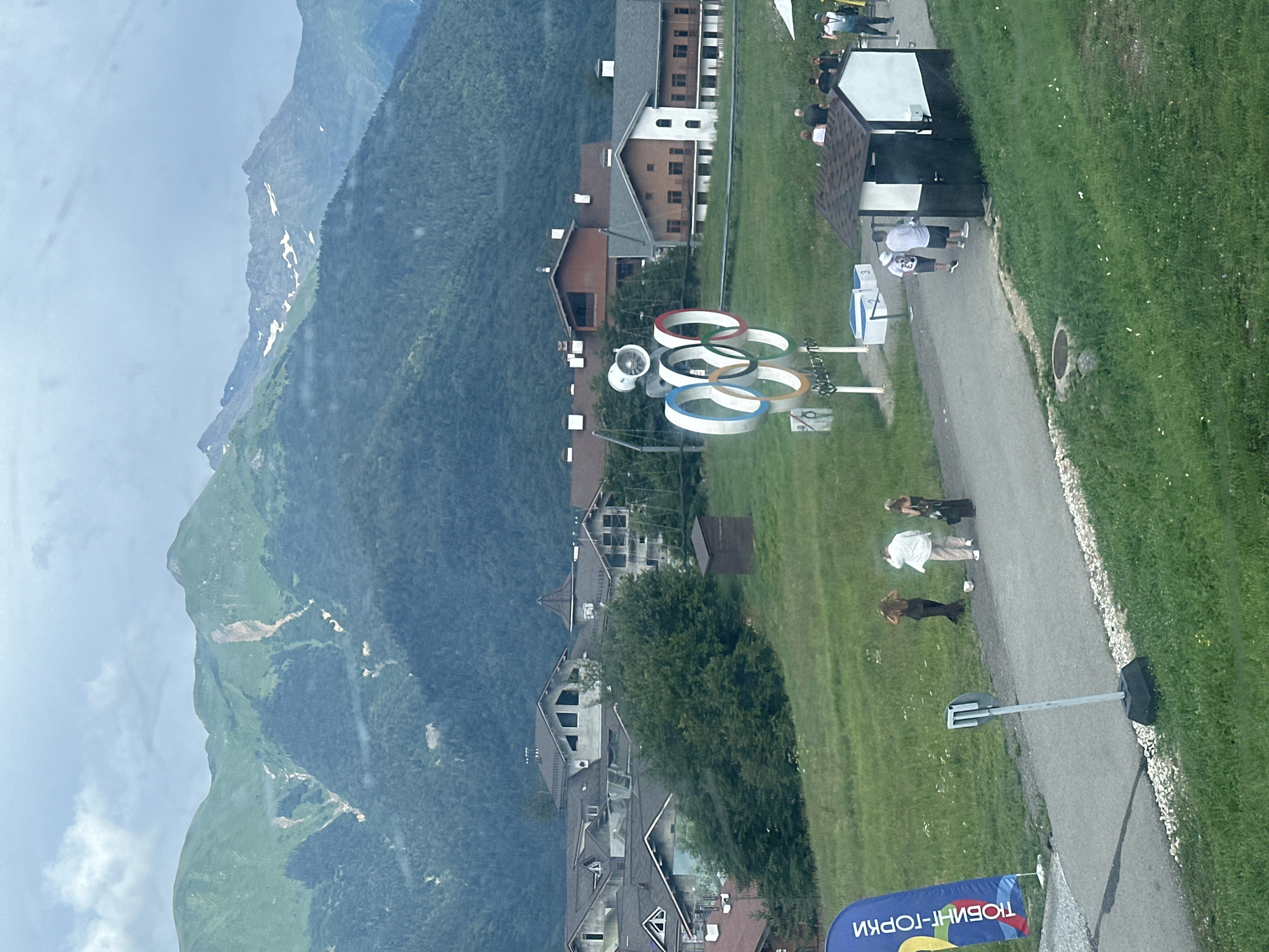 The image depicts a mountain landscape with a cluster of buildings in the mid-ground and a pathway with several people. In the background, a series of green, steep mountains dominate the horizon, with some patches of snow visible near the peaks. The mountains show signs of erosion and rockslides.
In the mid-ground, nestled amongst the trees, are several buildings with dark roofs and light-colored walls. Some of these buildings appear to be under construction, with scaffolding visible on one. Adjacent to the buildings, on a grassy slope, are the Olympic rings monument.
A paved pathway runs through the scene, with several individuals walking or standing. On the left side of the pathway, a blue banner with yellow text is displayed vertically. To the right of the Olympic rings, a small, dark-roofed structure with white walls is present. People are observed near this structure and further down the path. The ground is covered in green grass with scattered white wildflowers. The sky is overcast. The scene is likely located in Estosadok, Russia, a mountain resort known for hosting events during the 2014 Winter Olympics.