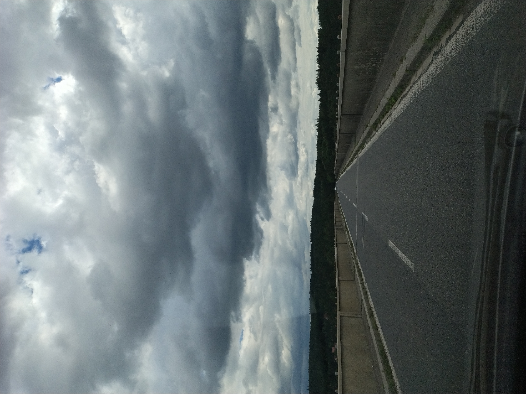 The image shows a view from a car driving on a highway bridge, likely in or near Podolí I, Czechia, on a cloudy day. The road is asphalt with a white line marking the lane. On either side of the road are concrete barriers topped with decorative spherical elements. Beyond the barriers, a dense green forest lines the horizon. The sky is filled with dramatic, dark gray clouds with patches of lighter gray and a small glimpse of blue sky. The overall impression is one of a slightly overcast and moody landscape.