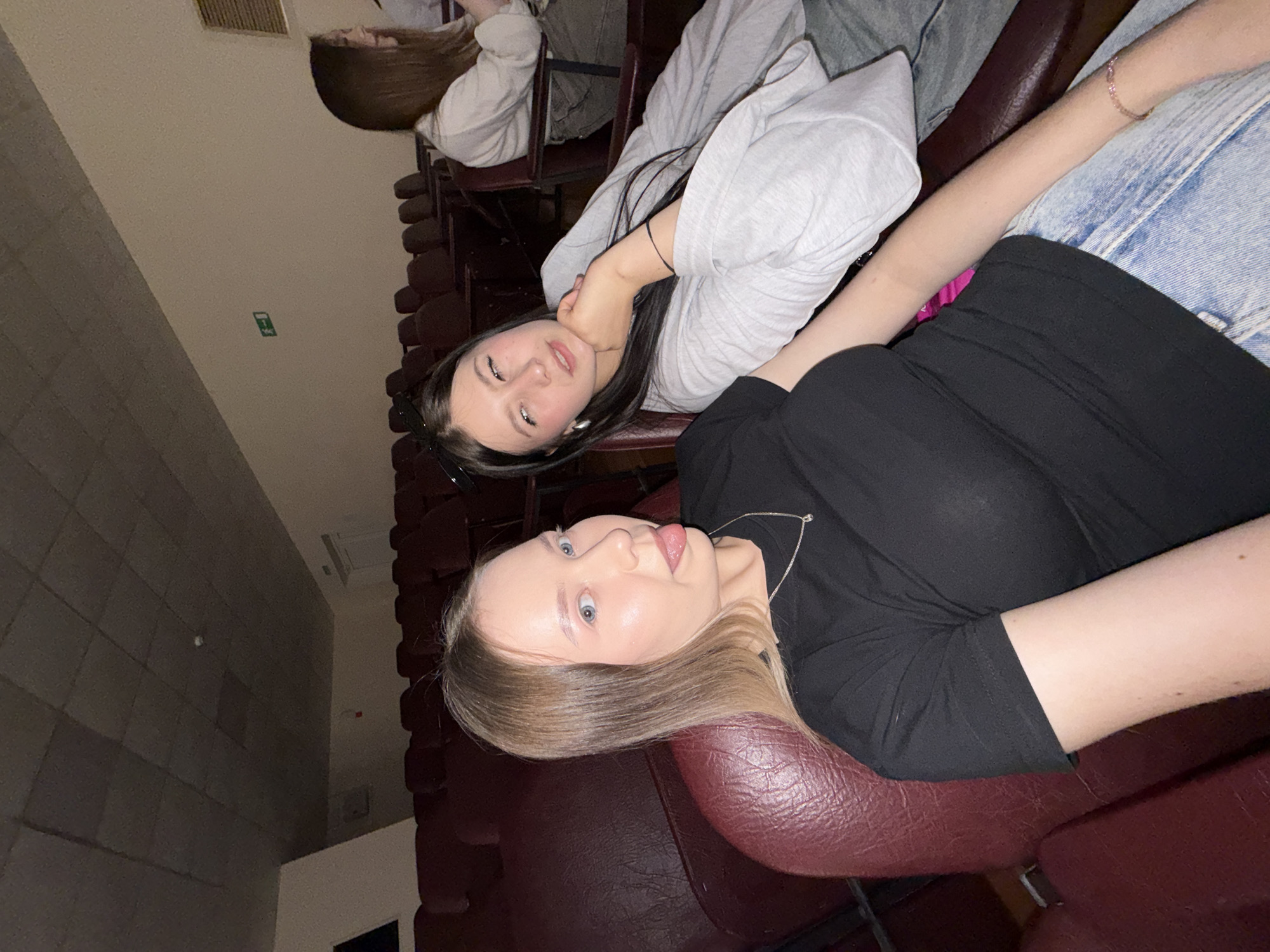 Two young women are lying in cinema seating. One woman has long dark hair and is wearing a grey sweatshirt and jeans. The other has blonde hair, is wearing a black t-shirt and jeans. There are several rows of unoccupied cinema seats visible. The location is likely a cinema in the city of Chaikovsky, Russia.