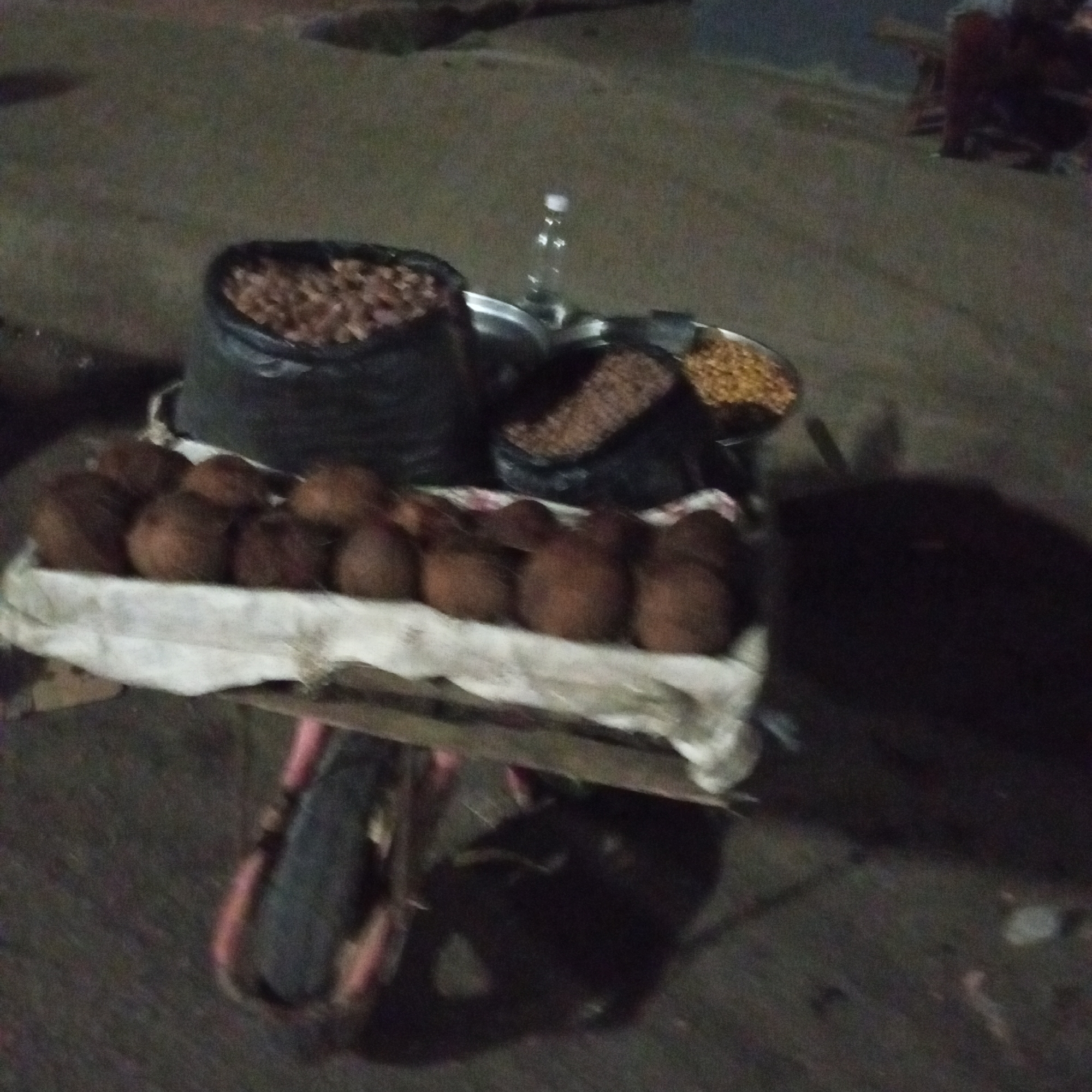 A street vendor's cart in Bukuru, Nigeria, displays coconuts, peanuts, and roasted groundnuts on a white cloth. A plastic bottle rests on a metal tray with additional nuts. The scene appears to be at night.