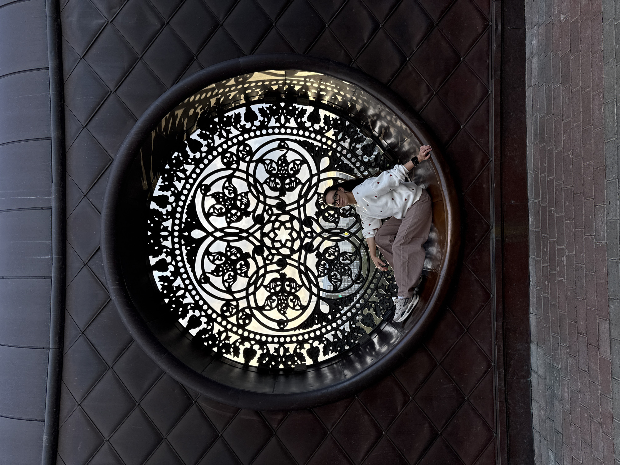 A woman is sitting in a large, circular, decorative window on a building facade in Kazan, Russia. The window features intricate metalwork with floral patterns. The building's facade is dark brown and has a quilted texture. The woman is wearing a white sweater with red heart patterns and brown pants. She is looking towards the camera with glasses on. The ground in front of the building is paved with brick.