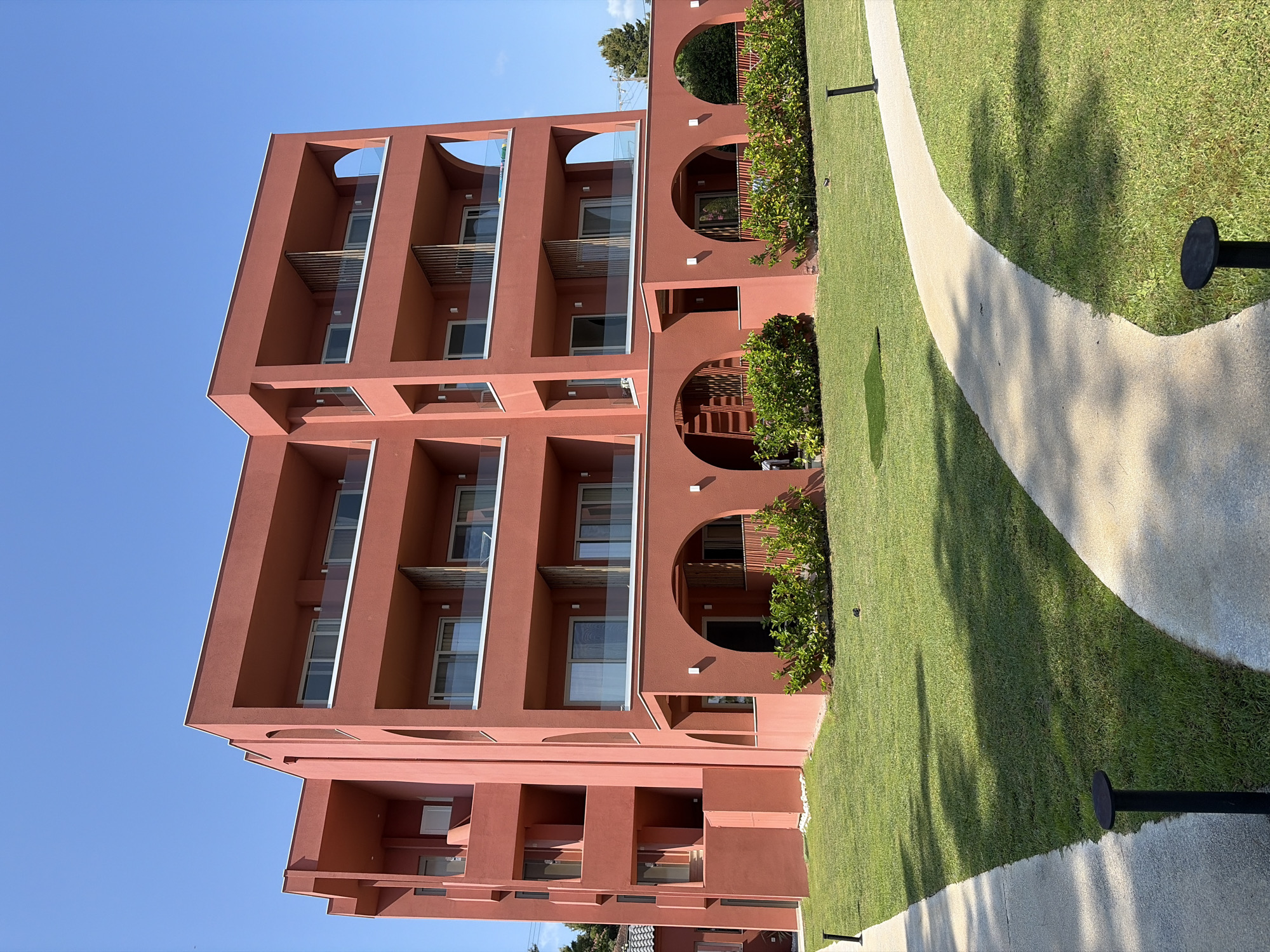 A modern, salmon-colored building with multiple balconies and arched ground-floor entrances stands under a clear blue sky. The building has a geometric design with recessed balconies and glass railings. In front of the building, a well-maintained green lawn is bordered by a curved concrete pathway. Several black, low-profile lampposts are spaced along the pathway. Lush green bushes are planted in front of the arched entrances on the ground level. The scene suggests a resort or hotel in a sunny, possibly coastal, location. This building is located in Pallini, Greece.