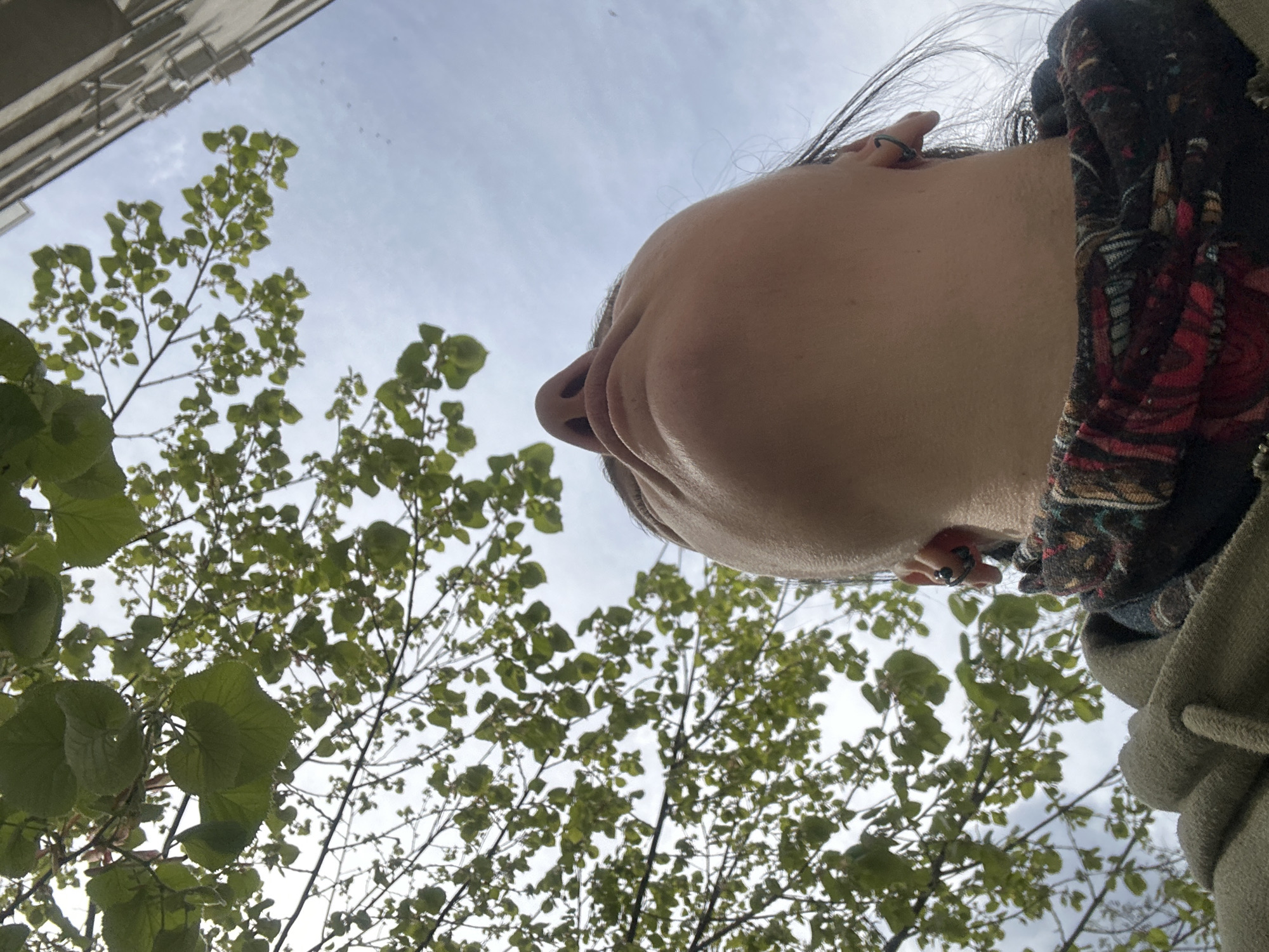 The image presents a unique worm's-eye view looking upwards at a person, trees, a building, and the sky in Sofia, Bulgaria.
The lower part of a person's face occupies the right side of the frame, seen from directly below. Visible features include their chin, nose, mouth, and the right side of their face, including an ear with a small, dark hoop-style earring. Dark hair is visible framing their head, and a patterned fabric, possibly a scarf or part of a garment with red, black, and other colors, is draped on their right side. A light-colored hoodie or similar casual clothing is also visible on their right shoulder.
Lush green foliage from several trees dominates the left and central portions of the image, with bright green leaves indicating springtime or summer. The trees' branches are silhouetted against the sky. In the upper left corner, a section of a tall, light-colored building with visible architectural details suggests an urban or suburban environment.
The sky is bright and appears light blue with scattered white clouds, indicating daylight conditions, possibly under soft or overcast light. The overall perspective suggests the person is looking directly upwards, perhaps observing the canopy of trees and the sky above. There is no visible text in the image.