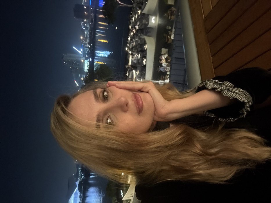 A woman with blonde wavy hair and green eyes rests her head in her hand. She wears a black top with ruffled sleeves. The background features a nighttime cityscape with illuminated buildings and a body of water reflecting the lights. Tables and chairs are visible in the distance, suggesting an outdoor dining area. The image is slightly blurred.