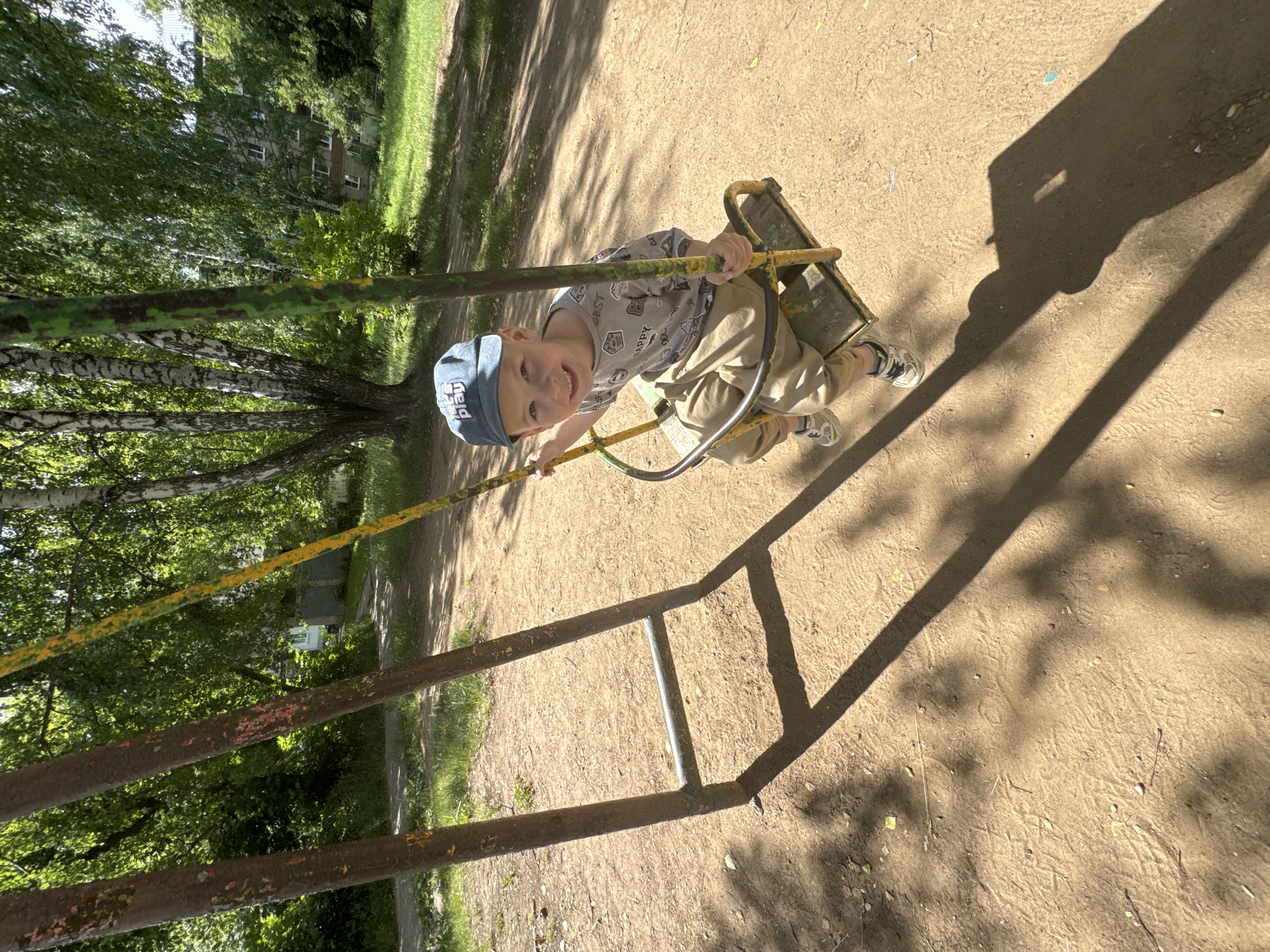 A young Caucasian boy with blond hair and blue eyes, wearing a blue baseball cap, a gray graphic t-shirt, and tan pants, is sitting on a yellow and green swing set. The boy is smiling and looking directly at the camera. He is holding onto the swing's chains. The swing set is located on a sandy playground. In the background, there are trees with green leaves, a building, and more trees. The scene appears to be outdoors on a sunny day, indicated by the shadows cast on the ground. The location is identified as the city of Chaikovsky, Russia.