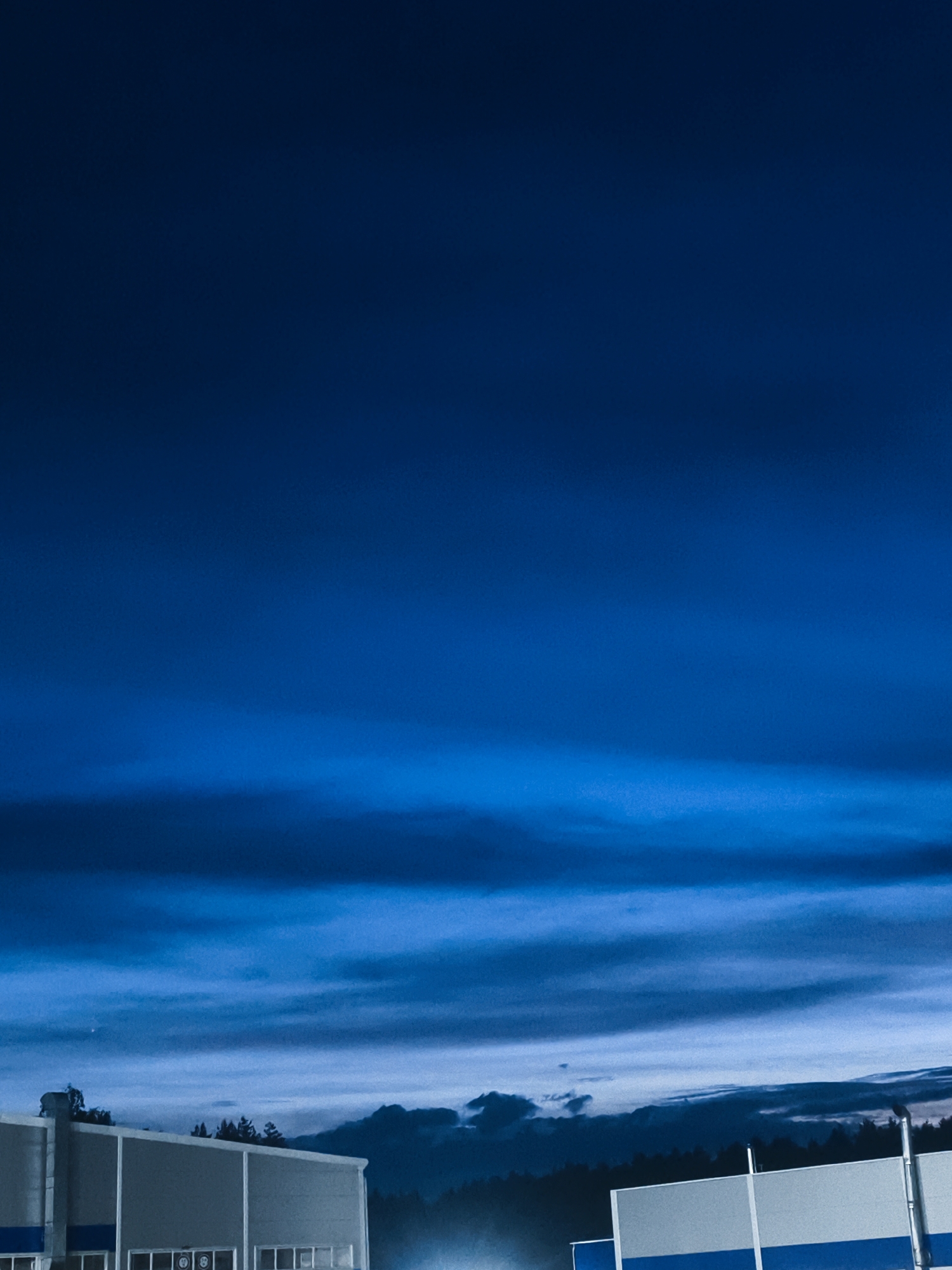 The image depicts a nighttime scene in what appears to be an industrial area. The sky is a deep blue with layered clouds. In the foreground are the silhouettes of buildings with flat roofs and visible architectural lines, possibly warehouses or factories. A dark line of trees is visible in the distance. Based on the prompt, this is possibly in Noviy, Russia. There is no visible human activity, and the environment appears quiet and still.