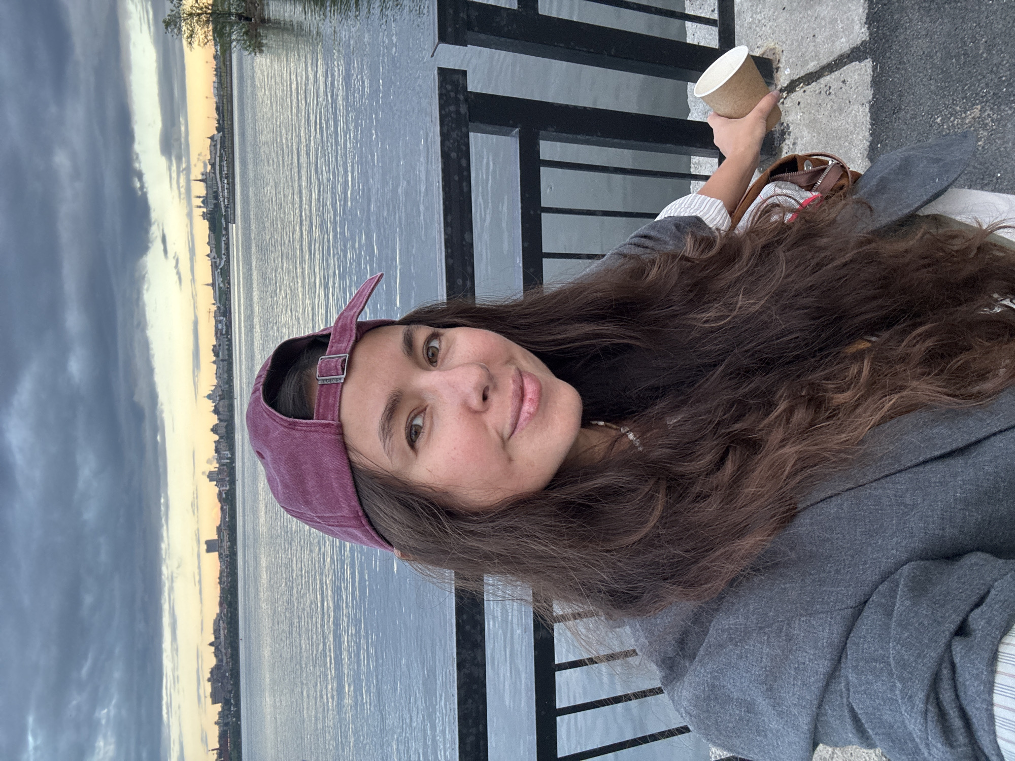 A woman wearing a maroon baseball cap and grey jacket holds a disposable cup while standing next to a black metal railing overlooking a body of water. The skyline of Kazan, Russia is visible across the water under a cloudy sky.