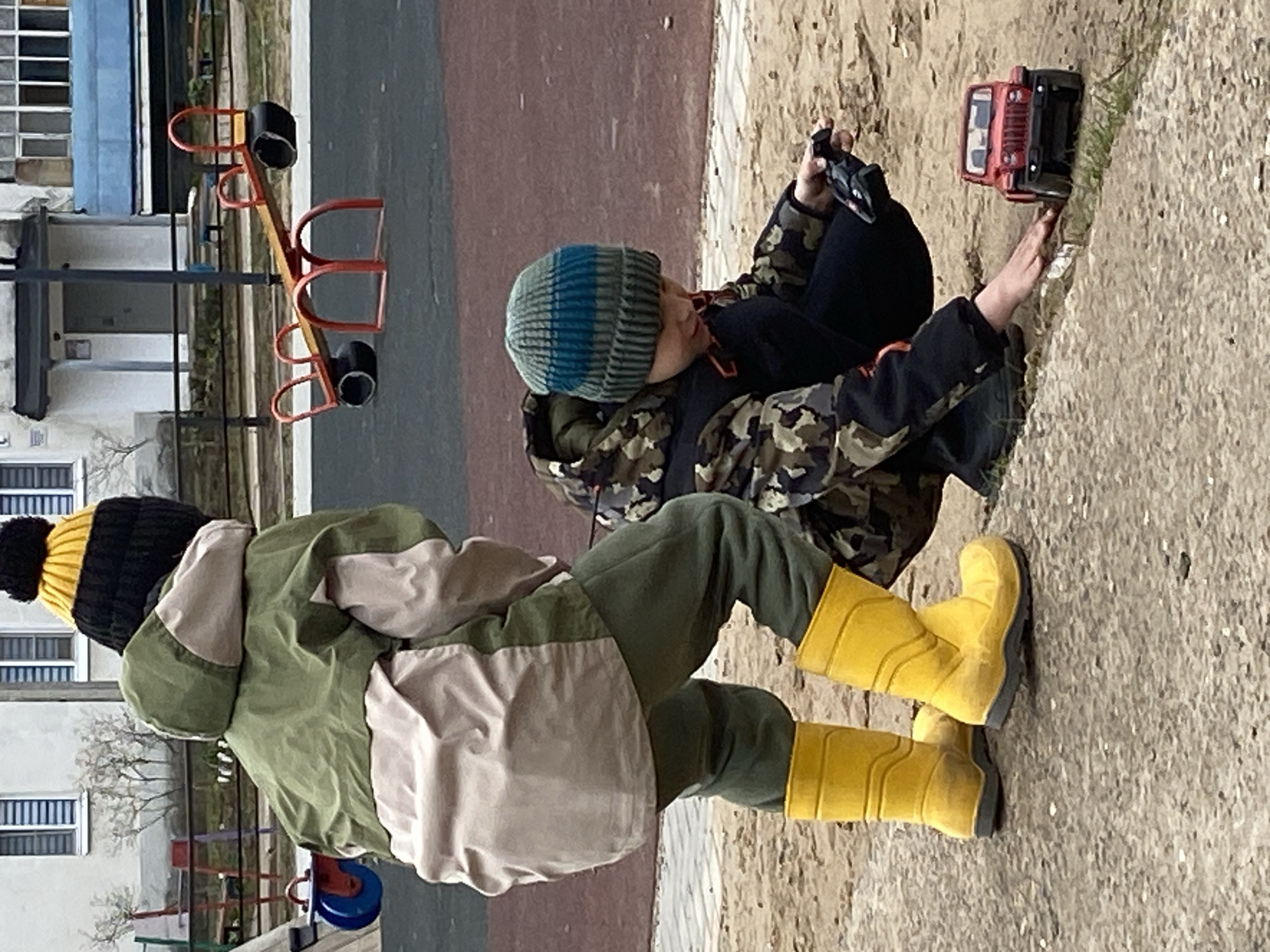 The image depicts two children playing outdoors in what appears to be a playground or courtyard setting in Noviy, Russia. The scene is set during the daytime, likely on a cool or cold day, given the children's warm attire.
One child is seated on a patch of sandy or dirt ground, wearing a camouflage-patterned jacket, dark pants, and a blue and grey striped knitted beanie. This child is actively engaged in playing with a red toy truck, holding a remote control device in their hands to operate it. The toy truck is positioned on the ground directly in front of the child.
Standing nearby is another child, viewed from behind, wearing a green and beige two-toned jacket, dark green pants, a black and yellow knitted beanie with a pom-pom, and bright yellow rubber boots. This child appears to be observing the play or waiting their turn.
In the background, a red and yellow seesaw is visible on a paved surface, characteristic of a playground. Beyond the playground area, residential buildings with multiple windows can be seen, suggesting an urban or suburban environment. The ground transitions from a dark paved or rubberized surface to a lighter, sandy or dirt area where the children are playing. The children seem focused and content in their activity. No discernible text is visible within the image.