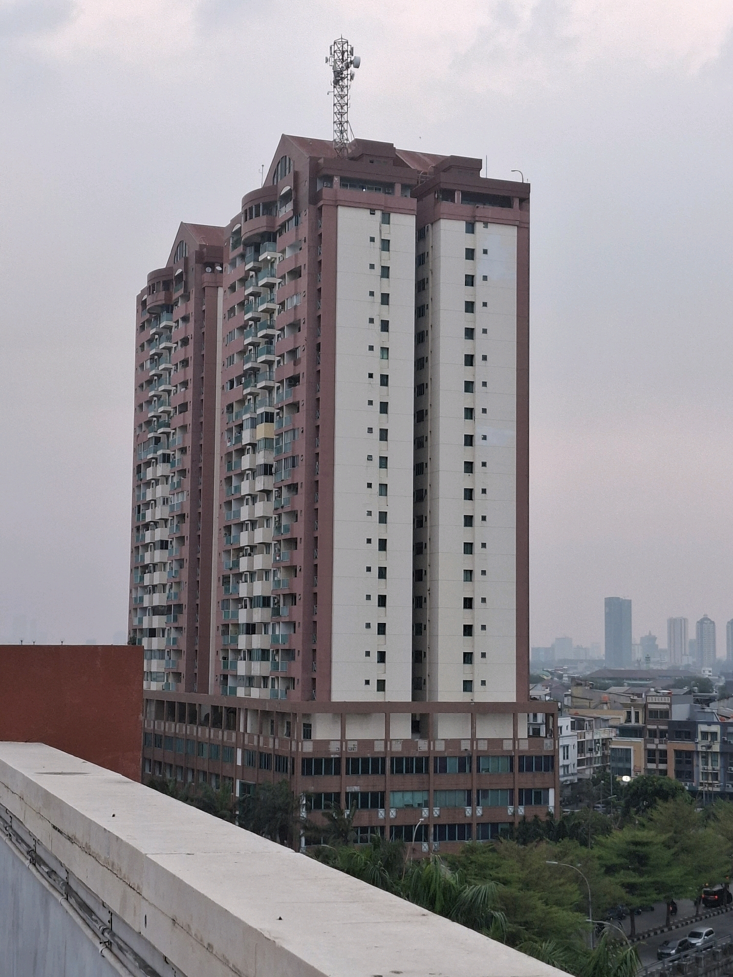 The image shows a large, multi-story residential building in a dense urban environment. The building has a distinctive reddish-brown and cream-colored facade with numerous balconies. A communication tower is visible on the rooftop of the building. In the background, other buildings and city structures can be seen, suggesting a developed urban landscape. The sky is overcast, and the overall atmosphere appears somewhat hazy, which is common in urban areas due to pollution or humidity. The foreground shows a concrete structure, possibly a rooftop or elevated walkway, with visible conduits and pipes. Trees and vegetation are present around the base of the building and in the surrounding cityscape, indicating some green spaces within the urban fabric. The presence of cars on a road in the lower right portion of the image further confirms the urban setting. Based on the architectural style and the overall urban density, this scene is likely located in a major city in Southeast Asia, such as Central Jakarta, Indonesia.