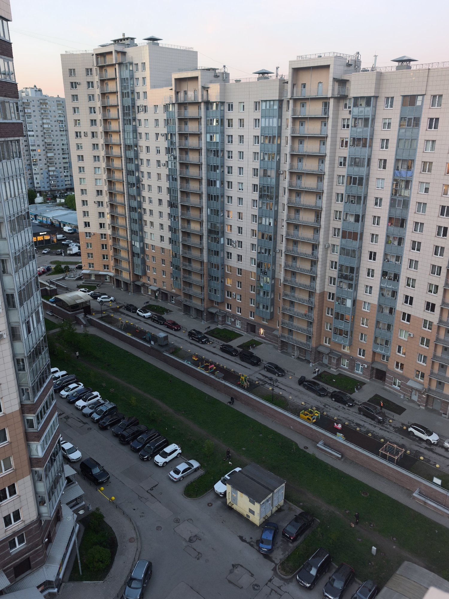 The image depicts a high-angle view of a residential area in Saint Petersburg, Russia. Multiple modern apartment buildings dominate the scene, characterized by their light-colored facades and numerous balconies. The buildings are arranged linearly along the frame. Below, a green strip of land separates two parking areas filled with various cars, predominantly dark in color. Several people are visible, some walking, some seemingly involved in construction or maintenance activities near the green strip. A small, yellow utility building or trailer sits adjacent to one of the parking areas. The lighting suggests either early morning or late evening. In the distance, additional apartment buildings are visible.