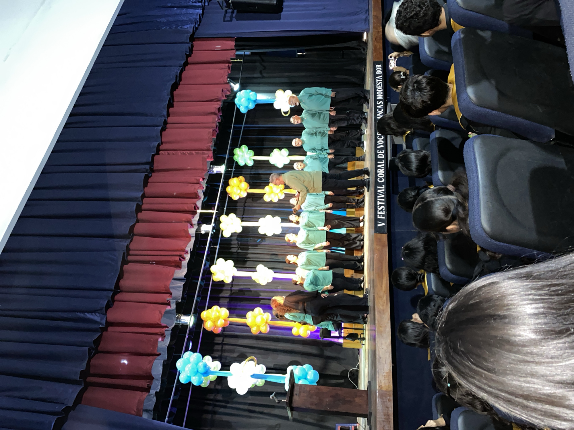 An indoor event, likely a choral festival, is captured from an audience perspective looking towards a stage. On stage, a group of approximately 10-12 adults, presumably a choir, are performing. Most are dressed in matching light green long-sleeved tops and dark trousers, standing in several rows. One individual in a light brown top and dark trousers is positioned forward, appearing to lead or conduct.
The stage features a wooden floor and a dark fabric backdrop. Decorative balloon arrangements, shaped like flowers or clouds in clusters of blue, white, yellow, and orange, stand on tall, thin poles across the stage. A wooden podium is visible on the far left of the stage. Dark blue and reddish-brown curtains frame the stage on the left.
The audience, composed of children and adults, occupies the foreground and right side of the image, seen from behind. They are seated in rows of blue auditorium-style chairs, attentively watching the performance. Several audience members are holding up smartphones, seemingly recording the event.
The setting appears to be an auditorium or school hall. The event is well-lit by artificial stage lighting. Partially visible text on a white banner or backdrop to the right of the stage reads "V FESTIVAL CORAL DE VOCES INFANTILES MODESTA BOR," identifying the event as the "V Choral Festival of Children's Voices Modesta Bor."
