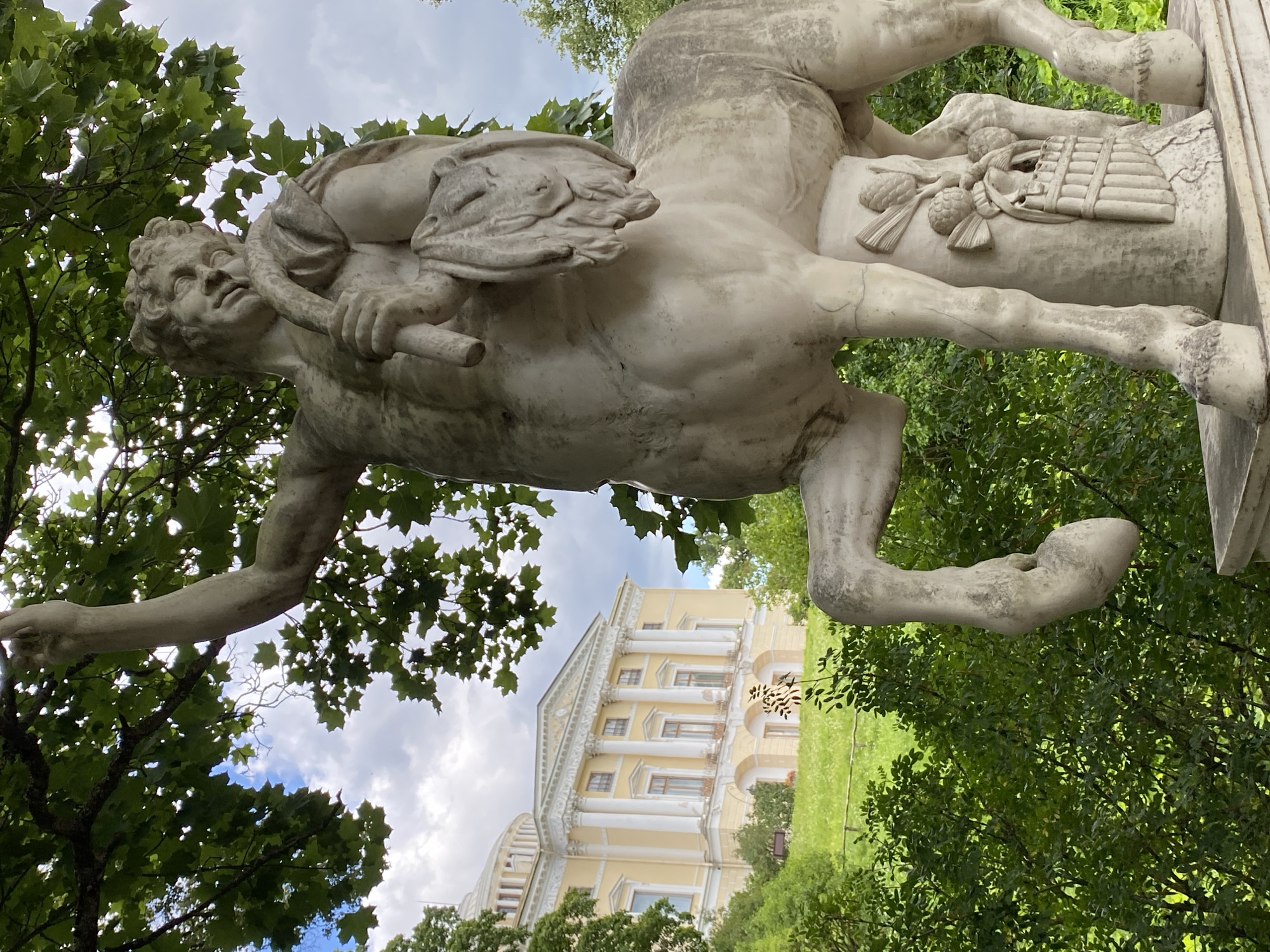 The image shows a marble sculpture of a centaur in the foreground, partially obscured by trees. The centaur is depicted with a human torso attached to the body of a horse. It is white in color and appears weathered. The sculpture stands on a pedestal with relief carvings of pinecones and a pan flute. In the background, a large, neoclassical yellow building with white columns and a dome is visible, situated on a grassy hill. Lush green trees and foliage surround the sculpture and the building. The sky is partially cloudy. This scene is located in Pavlovsk Park, Pavlovsk, Russia.