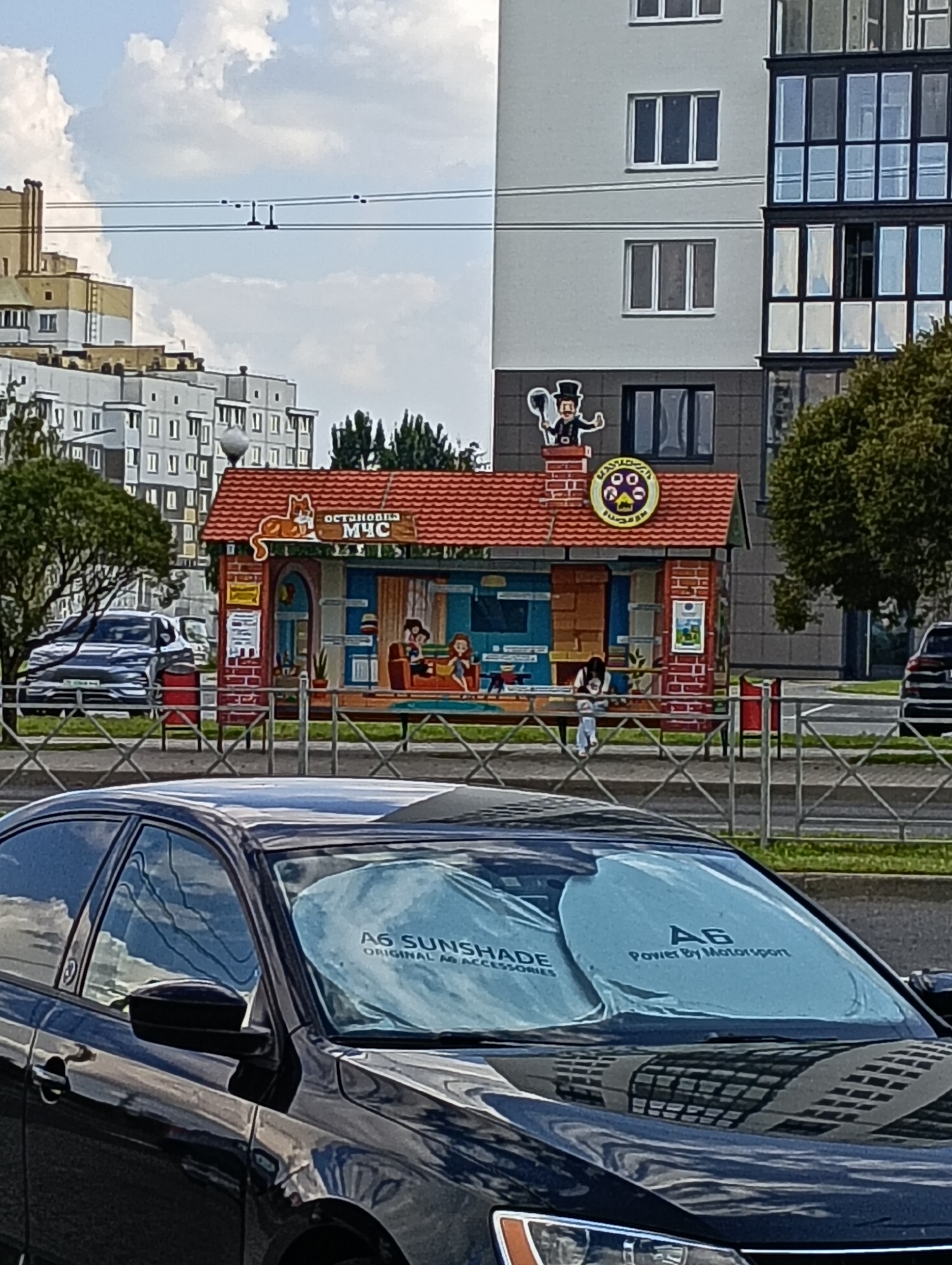 In Hrodna, Belarus, a picturesque scene unfolds with a whimsical kiosk as the focal point. The kiosk, resembling a miniature house, is adorned with a vibrant red, tiled roof, and its facade is decorated with colorful murals depicting a cat on the roof and stylized figures inside. Above the kiosk, a chimney sports a chimney sweep character, adding to the charm. The surrounding environment includes a modern residential building with numerous windows, suggesting an urban setting. Power lines crisscross the sky, a common sight in many cities. In the foreground, a black car is parked, its windshield covered by a sunshade with the inscription "A6 SUNSHADE ORIGINAL A6 ACCESSORIES POWER BY MOTORSPORT". The overall impression is one of a lively, everyday urban environment with a touch of playful artistry.