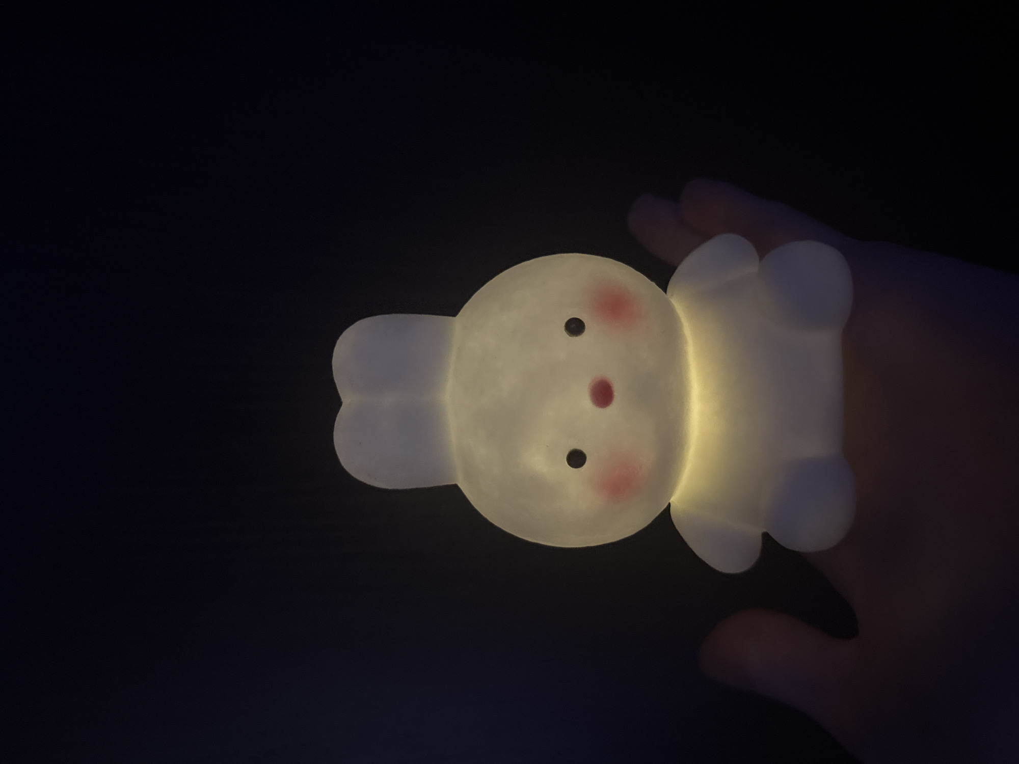 A cute, glowing, white bunny-shaped lamp is being held in a person's hand against a black background. The bunny has two large ears pointing upwards, black dot eyes, rosy cheeks, and a small pink nose. The lamp emits a warm, yellow light from its lower half. The hand holding the lamp is dimly lit and appears to be a person's palm with fingers curled around the base of the lamp. The scene suggests a nighttime or dimly lit indoor environment, as the background is entirely dark. There are no discernible landmarks or environmental clues to indicate a specific city or location.