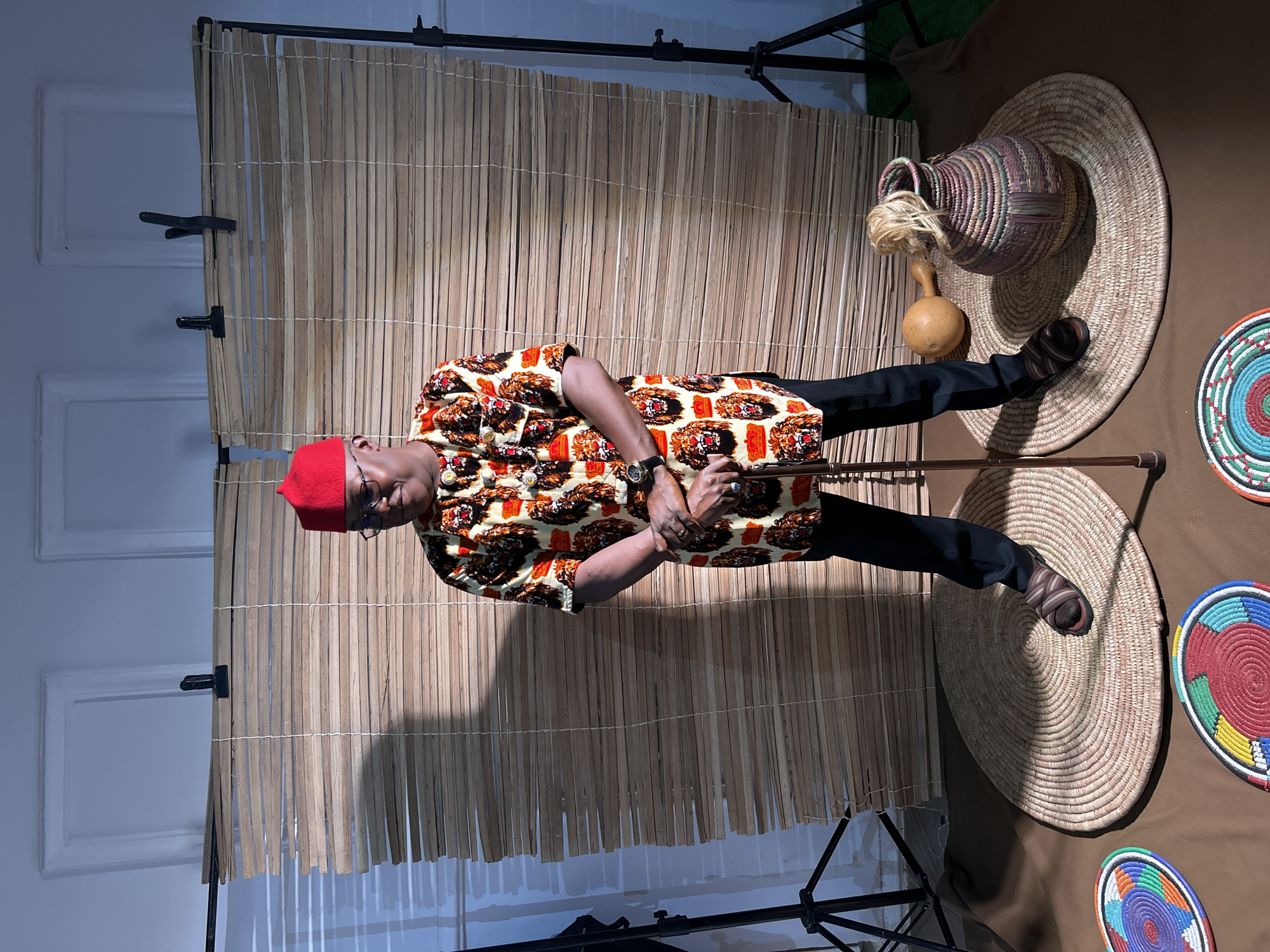 A man stands in front of a bamboo screen in a studio setting. He wears a patterned shirt, black pants, glasses, and a red hat. He holds a cane in his hands. To his right, there are woven mats, a woven basket, and a calabash gourd. The scene is likely a staged photograph, though the location (Jos, Nigeria) is not apparent from the visual information alone. A light stand is visible in the foreground.