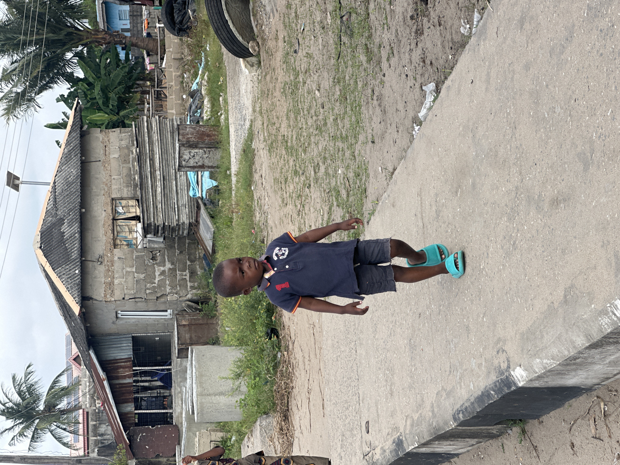 In Badagry, Nigeria, a young boy walks on a concrete path beside a road. He wears a dark blue polo shirt with an orange trim and the number '1' on the chest, along with dark grey shorts and bright turquoise sandals. In the background, a building constructed with grey concrete blocks has a corrugated metal roof. To the left of the building, a palm tree stands tall. The ground is a mixture of dirt and sparse green grass, with debris scattered about, including a large black tire and a rusted metal barrel. The sky is overcast.