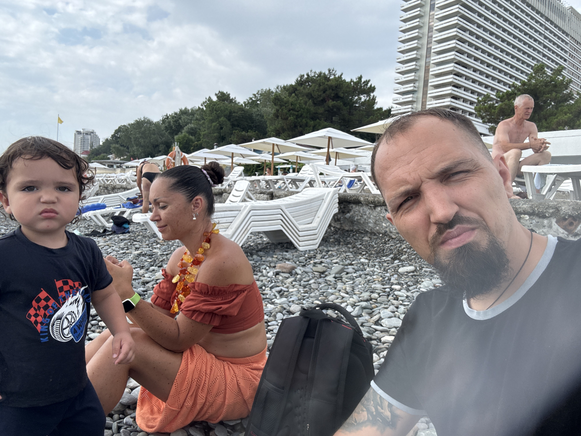 A family is on a pebble beach in Sochi, Russia, near a large, modern hotel building. In the foreground, a young child in a black t-shirt with a checkered flag and car graphic stands to the left, looking at the camera with a serious expression. To the child's right, a woman wearing an orange off-the-shoulder top and a matching orange skirt with an intricate pattern is seated, looking down. She has a prominent multi-colored beaded necklace. A black backpack rests on the pebbles behind her. In the middle ground, several white lounge chairs are stacked, and umbrellas are set up. Further in the background, a shirtless older man with gray hair is seated on a lounge chair, looking to his left. The sky is overcast.