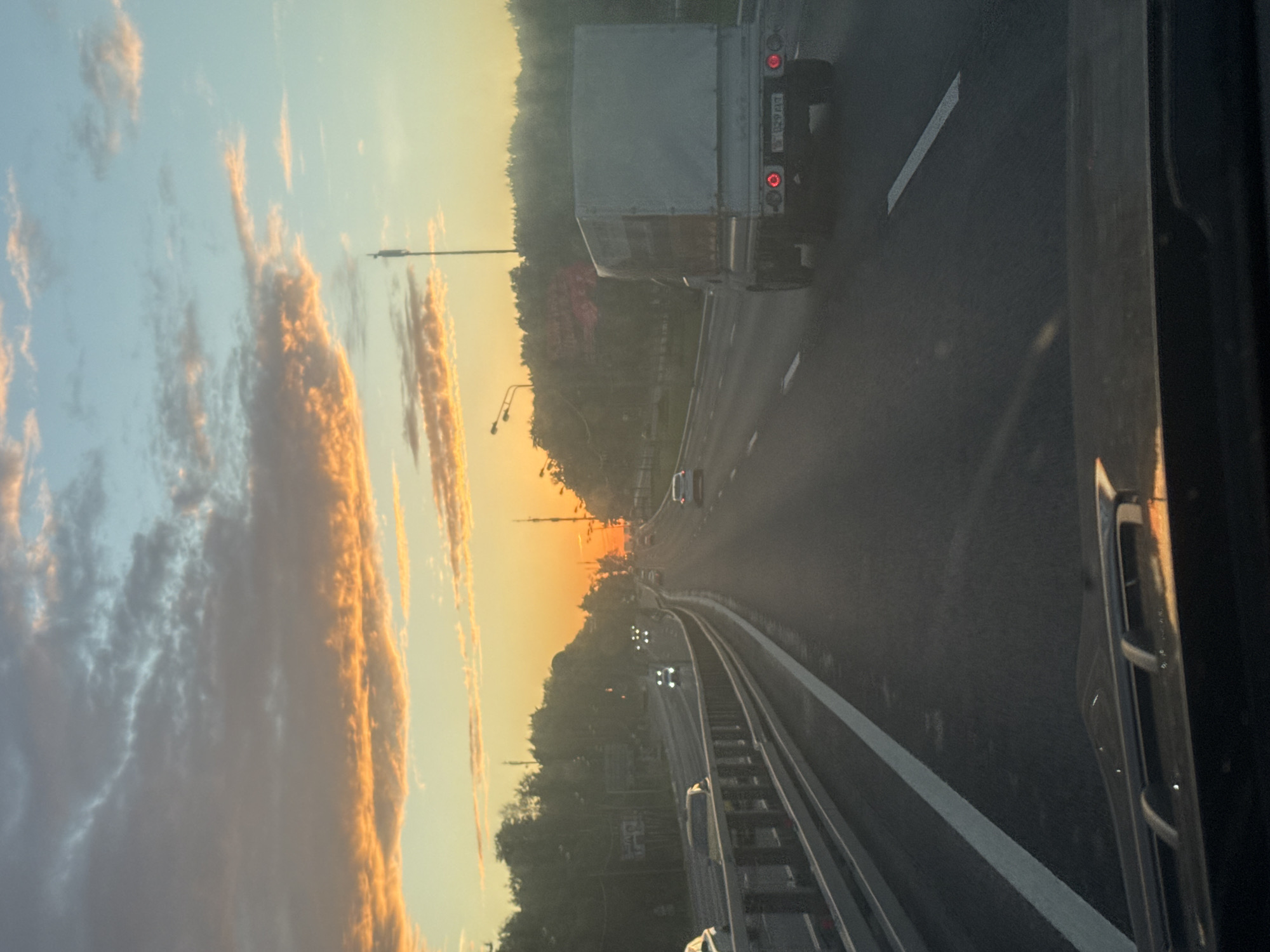 This image captures a sunset scene on a highway, likely in Moscow, Russia. The perspective is from inside a vehicle, looking forward and slightly to the left. The sky is a vibrant spectacle of sunset colors, with streaks of orange and yellow clouds against a pale blue backdrop.
In the foreground, a truck with a white box trailer is visible in the right lane of the highway, its taillights illuminated. The highway itself has clear lane markings and a barrier separating it from what appears to be a parallel railway track or another elevated section. Further down the road, other vehicles are present, some with their headlights on, indicating the waning daylight.
The roadside is lined with trees, and streetlights can be seen in the distance. The setting sun is casting a warm glow over the landscape, creating a serene and slightly hazy atmosphere. The overall mood is one of travel and transition, perhaps a journey home or to a destination as the day ends. No people are clearly discernible, but the presence of vehicles implies human activity. The time of day is clearly sunset due to the lighting and cloud colors. The location is hinted at by the mention of Moscow, Russia. No discernible text is visible in the image.