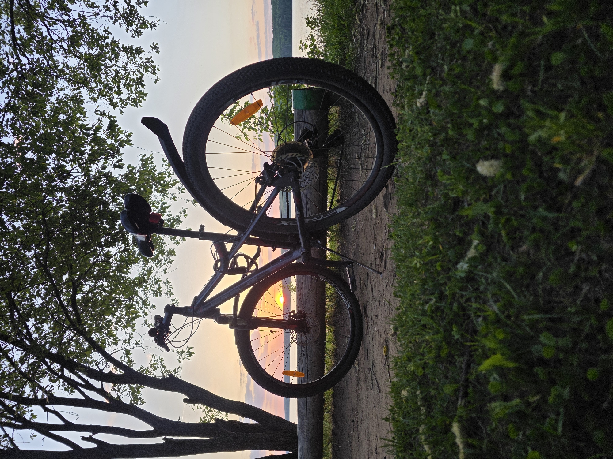 A bicycle is parked on a wooden fence post with vegetation to the right. Silhouetted tree branches are to the left. The sunset on the Kama River is in the background. The location is likely in the city of Chaikovsky, Russia based on the image context.