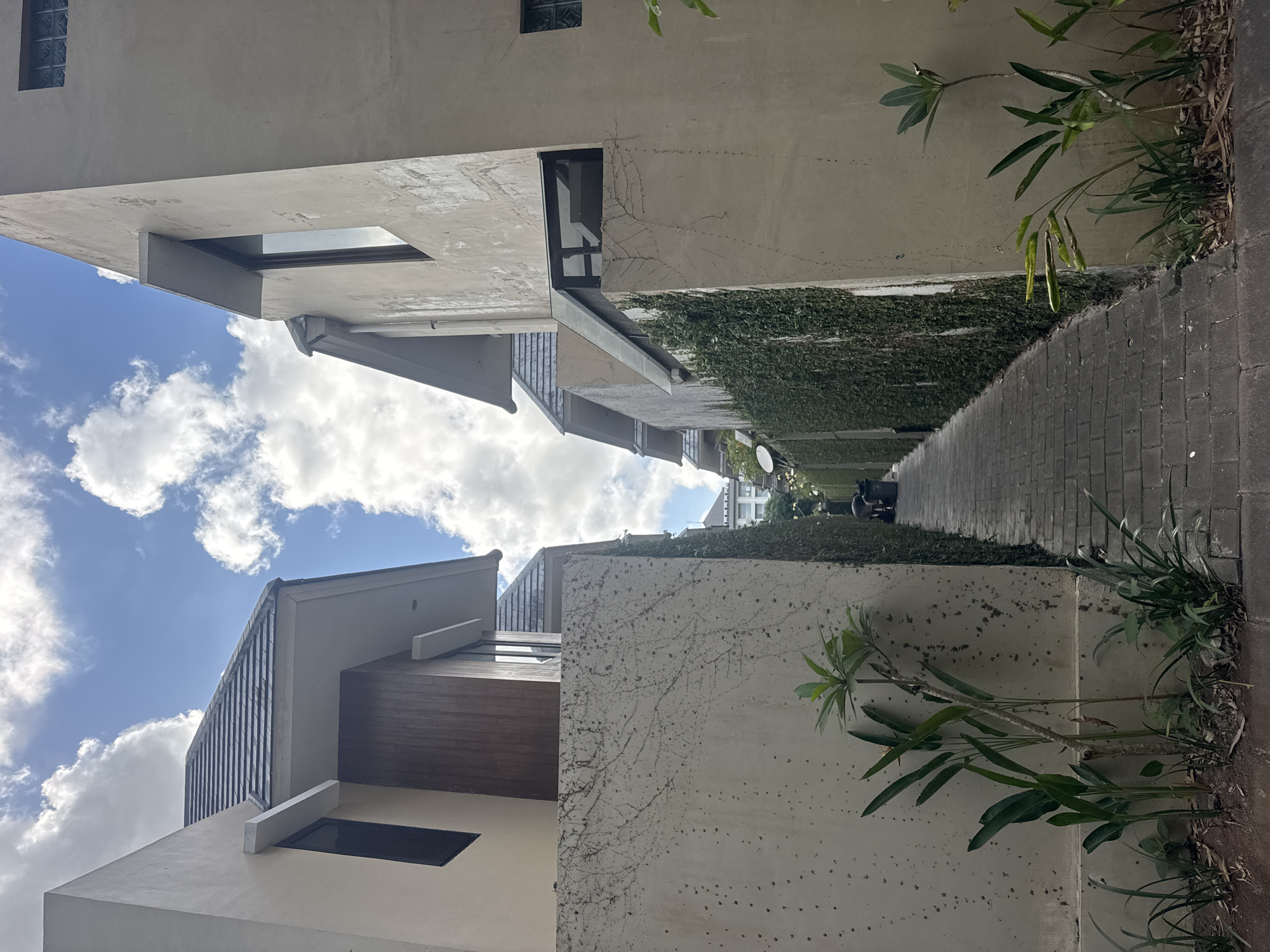 The image shows a narrow pathway between two modern buildings, likely in the city of Kuta Selatan, Indonesia. The pathway is paved with dark grey brickwork. Both buildings have light-colored, textured walls. The building on the left has a dark-tiled, sloped roof with a wooden accent on its upper facade. The building on the right has a flat roof and a large window with a dark frame. Green ivy covers the wall of the building on the right. Several small, leafy plants are growing along the base of the walls on both sides of the pathway. In the distance, at the end of the pathway, two large black trash bins are visible next to a person who is bent over, facing away from the camera, possibly interacting with the bins. The sky above is blue with scattered white clouds.