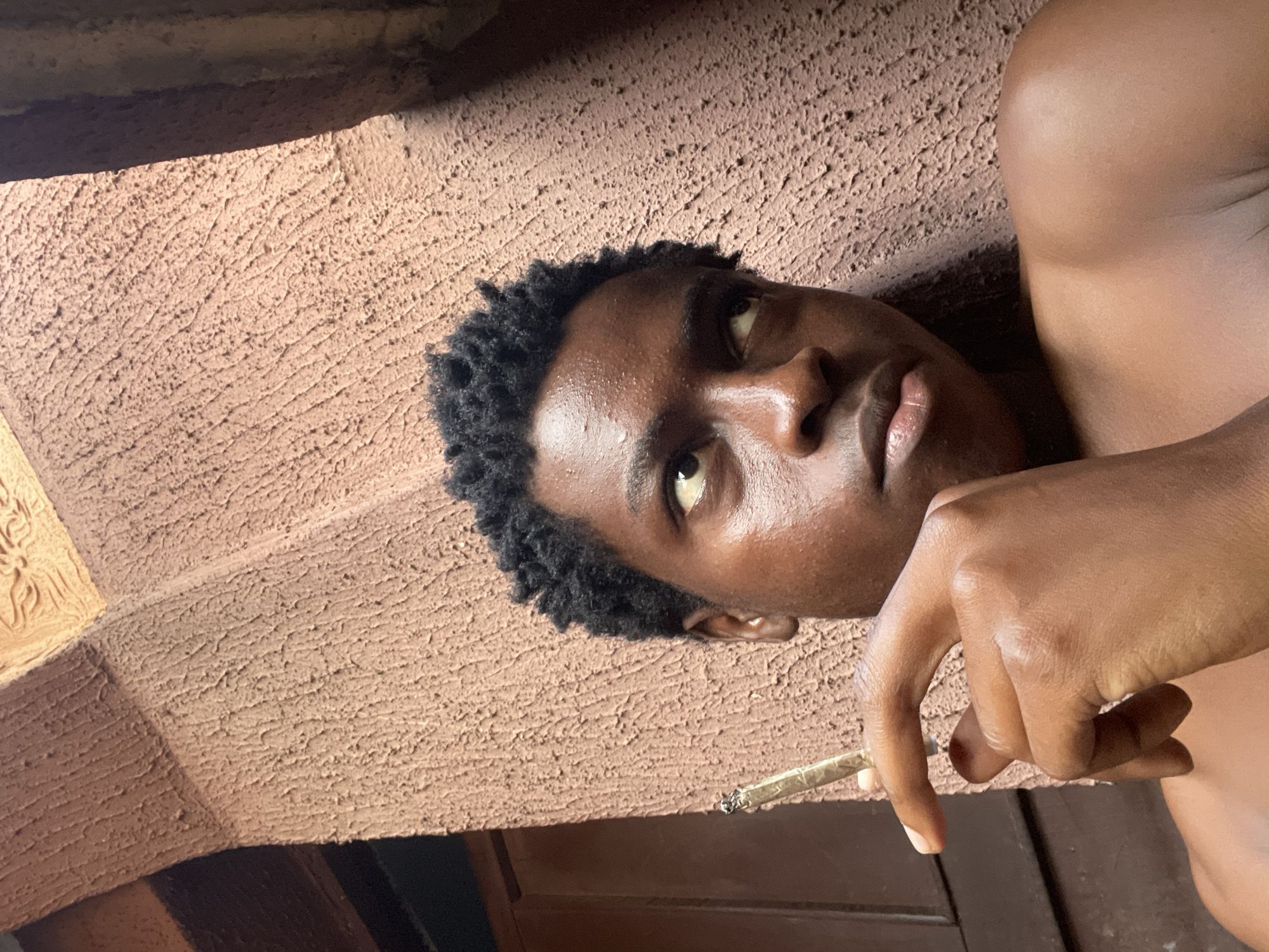 The image shows a close-up, low-angle shot of a young Black person lying down, looking upwards. The person has dark, curly hair and is holding a rolled cigarette between their fingers. Their skin appears to be tanned and slightly sweaty, suggesting it might be a warm day. The background is a textured, reddish-brown surface, possibly a wall or a rough-hewn material. There's a shadow cast across the upper part of the image, indicating sunlight, but the overall lighting suggests it could be late afternoon or early morning. The person's expression is ambiguous, a mix of contemplation and relaxation. The setting appears to be outdoors, given the natural light and the rough, possibly weathered texture of the background. There is no visible text in the image. The context provided indicates the location is Nkpor, Nigeria.