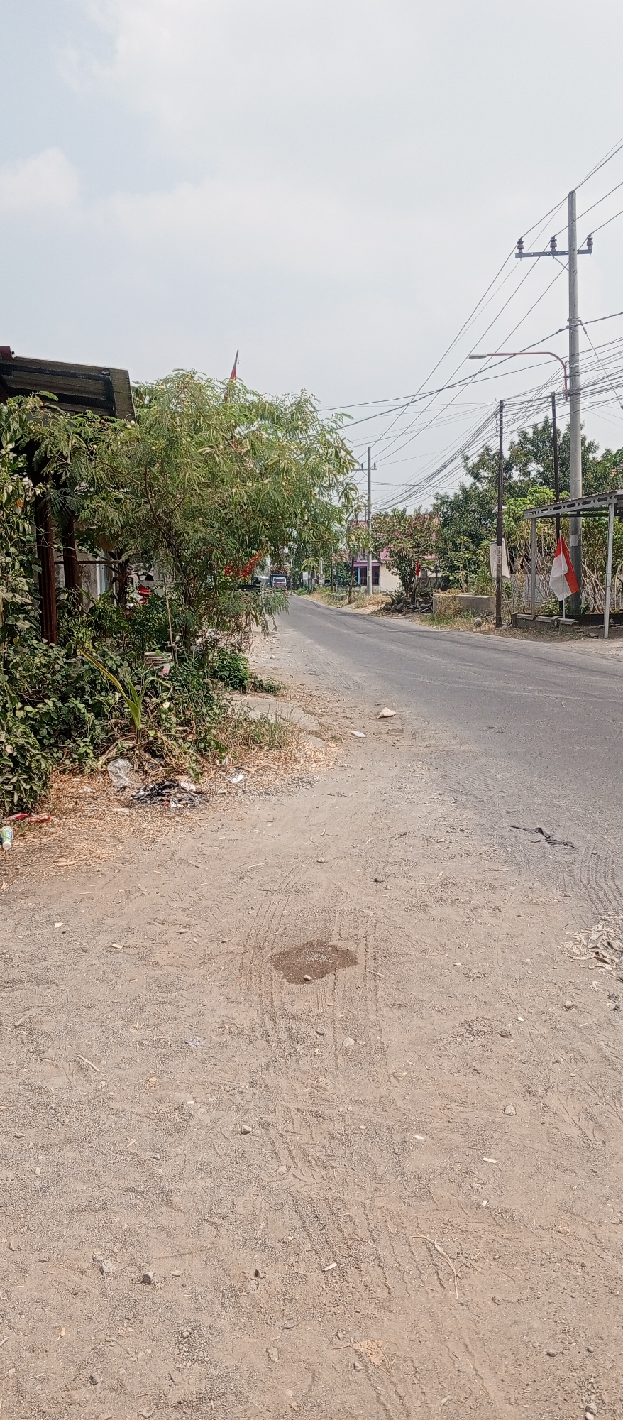 The image depicts a sunlit outdoor scene on a street in Gulun, Indonesia. A paved road occupies the right side of the frame, curving slightly into the distance. The road is flanked by a dirt shoulder on the left, littered with some debris, including plastic bottles and what appears to be burnt material. A large tree with abundant green foliage grows on the dirt shoulder, casting a shadow. To the left of the tree, a structure with a corrugated metal roof is partially visible. Several utility poles with a complex network of wires are present along the roadside, particularly on the right side, where a street lamp is also attached to one of the poles. In the background, several buildings with red roofs are visible, interspersed with trees. A small Indonesian flag, red and white, is displayed on a pole near a building on the right side of the road. The sky is overcast with scattered clouds. The overall impression is that of a residential or semi-rural area on a bright day.