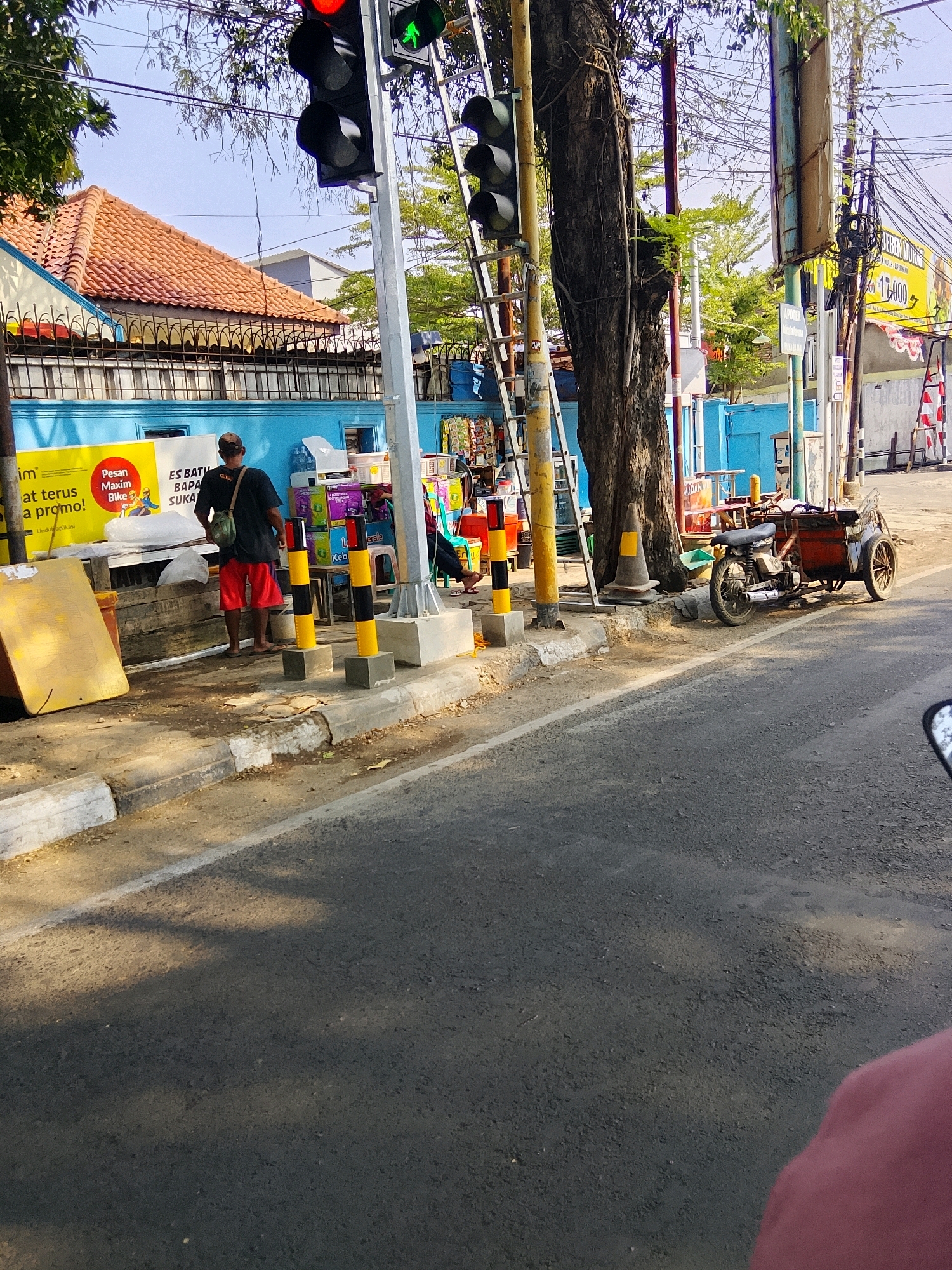A street scene in Cirebon, Indonesia, shows a man in red shorts and a dark shirt standing by a roadside stall. Traffic lights are visible, with one showing green. A motorbike with a cart is parked nearby. The stall appears to be selling various items, including drinks and snacks. The street is paved with asphalt, and there is a sidewalk with a curb. Trees and power lines are also present in the background.