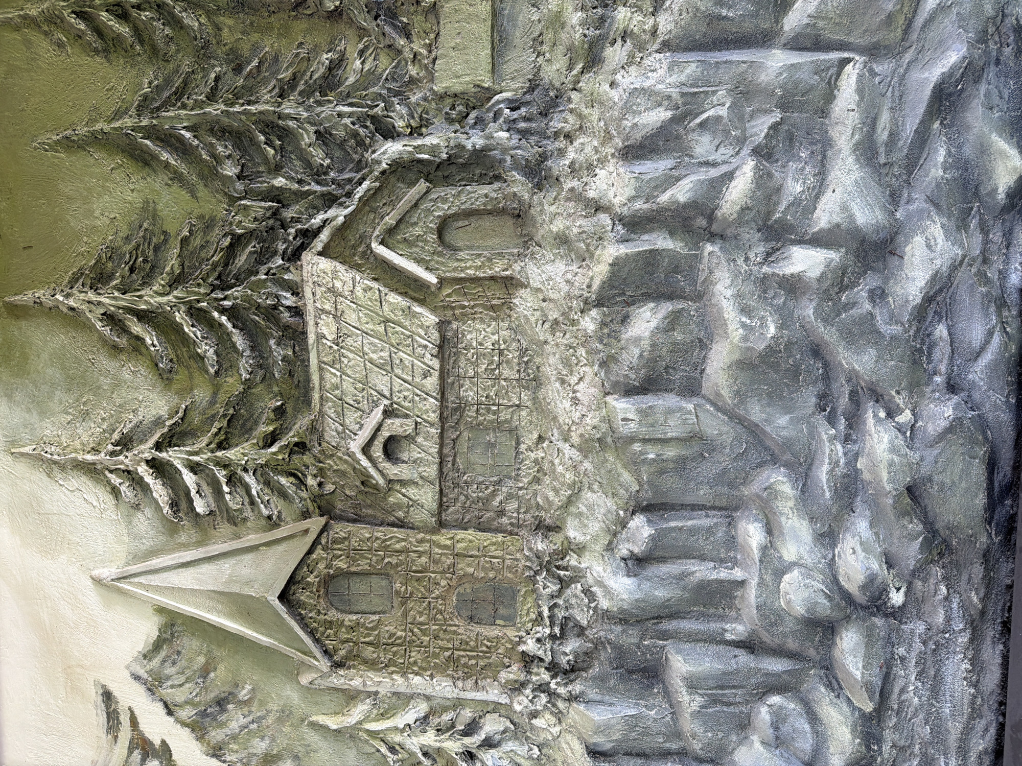 This is a close-up, top-down, eye-level, medium shot of a textured relief artwork depicting a mountainous scene with a village. The artwork is rendered in muted greens, grays, and beige tones. In the foreground, there is a rocky cliff face with a rough, uneven texture, appearing to be made of sculpted stone.
Dominating the mid-ground are two structures. To the left, a building with a tall, pointed steeple resembles a church or a tower, constructed with a stone-like pattern on its walls. It features arched windows. Adjacent to it, to the right, is a smaller, house-like structure with a gabled roof, also textured to appear like stone. This building has a small, arched doorway and a window.
Behind the structures, a cluster of evergreen trees with a distinct, textured appearance rises upwards. The trees are depicted with dark trunks and needle-like foliage, some of which appear to be dusted with a light color, possibly representing snow. The background is a textured expanse of a lighter, muted color, with faint suggestions of distant mountains or sky.
The artwork is highly textured, with deep grooves and raised areas that create a strong sense of depth and dimension. The overall impression is a rustic, mountainous village set against a backdrop of evergreen forest. There is no indication of the city of Bac Nha Trang Ward, Vietnam, within the visual elements of the artwork.