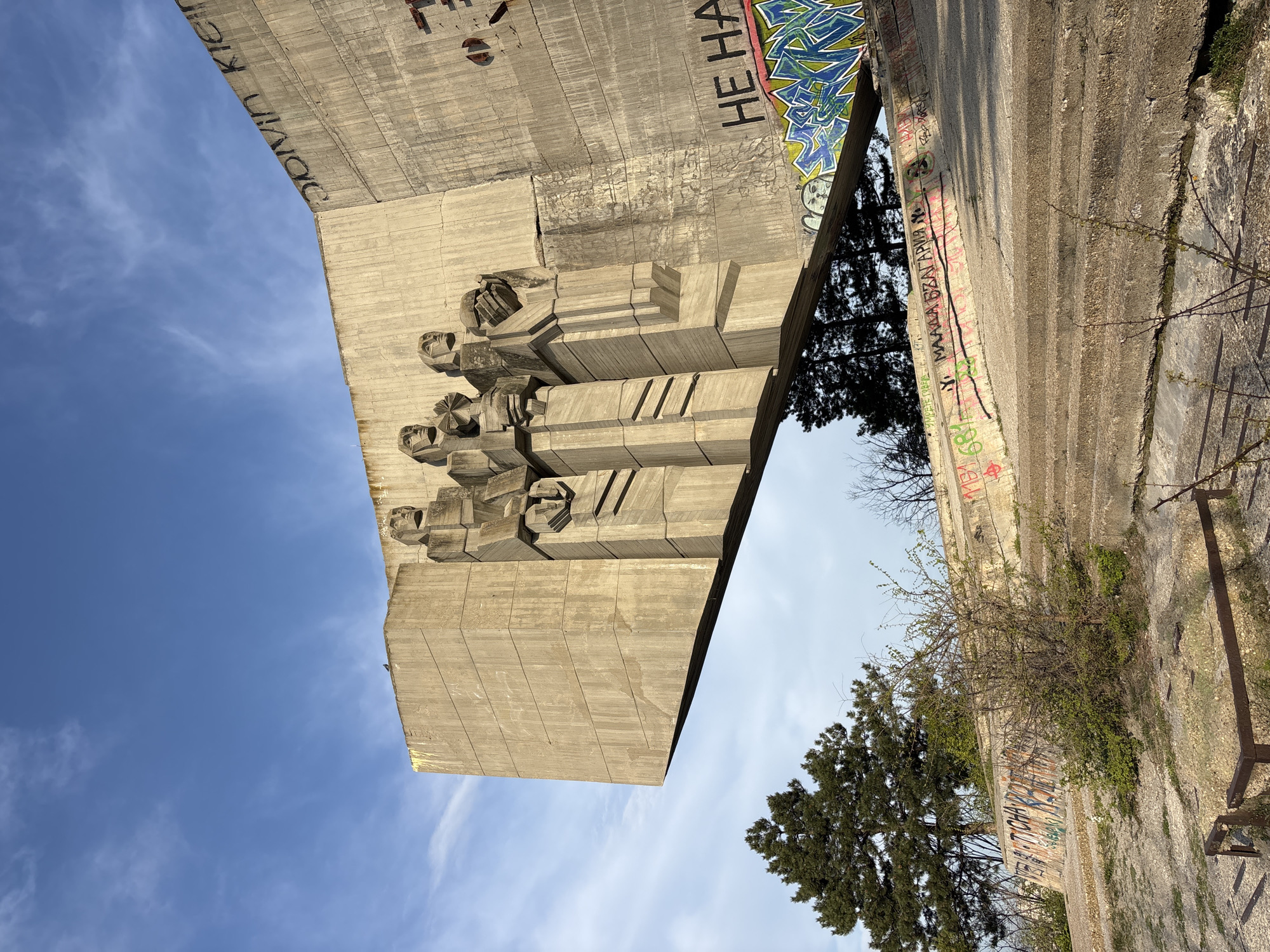 A low-angle shot shows a large concrete monument against a blue sky. The monument is adorned with blocky, geometric figures resembling human faces and abstract shapes. Graffiti marks the monument, some obscured by tree foliage. A concrete path or wall runs alongside the monument, also marked with graffiti and showing signs of vegetation growth. A few trees dot the landscape. The monument's details suggest a brutalist architectural style.