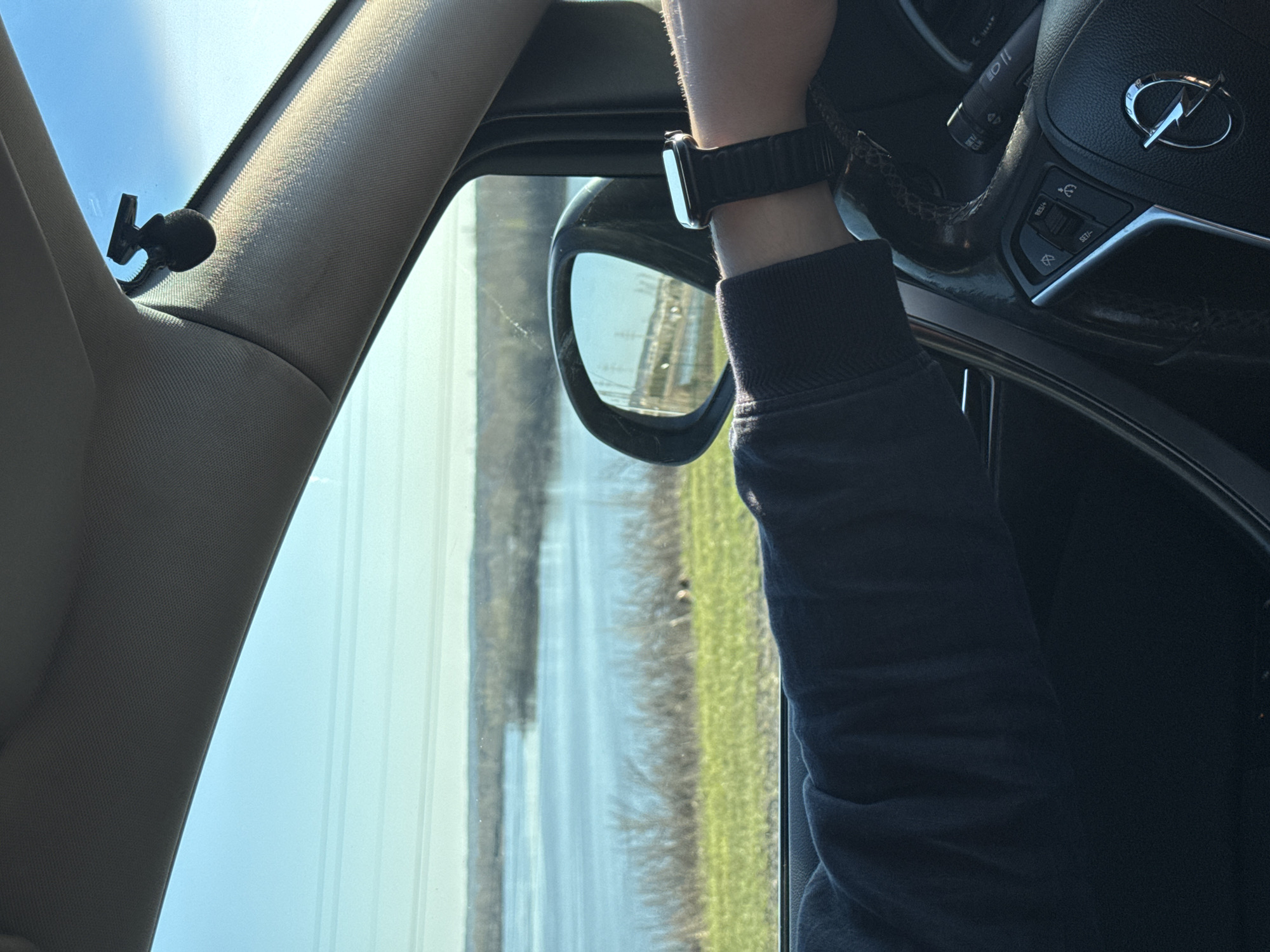The image depicts the interior of a vehicle, likely a car, with a focus on the driver's side. The driver's left arm is visible, wearing a black smartwatch with a black strap. The driver is holding the steering wheel, which features the Opel logo. The side mirror is visible, reflecting a landscape with a waterway and what appears to be green fields or vegetation. Outside, power lines are visible. The scene is shot during daylight hours, based on the brightness. The specific location in the city of Chaikovsky, Russia, cannot be determined from the provided information.