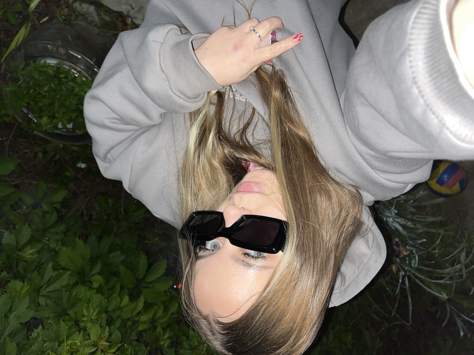 A woman with blond hair and wearing black sunglasses and a gray hoodie is pictured outdoors, with greenery visible at the bottom left and what appears to be a volleyball to the right of her head. She is holding her hand up, showing a ring and red nail polish. The image is taken from an elevated angle, slightly askew. In the upper left corner, there's a bucket filled with green plants. The location is Novopokrovskaya, Russia.
