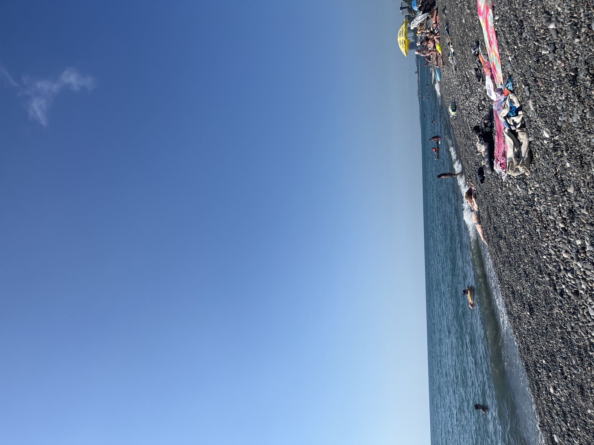 The image depicts a sunny day at a pebble beach in Kobuleti, Georgia. The sky is a clear blue with a few wispy clouds. The sea is a deep blue, with gentle waves lapping at the shore. Numerous people are enjoying the beach. Some are swimming in the sea, while others are sunbathing on towels and loungers on the pebbled shore. A yellow umbrella provides shade for a group of people. There are also several towels and clothing items scattered on the beach. In the background, buildings of Kobuleti are visible along the coastline.