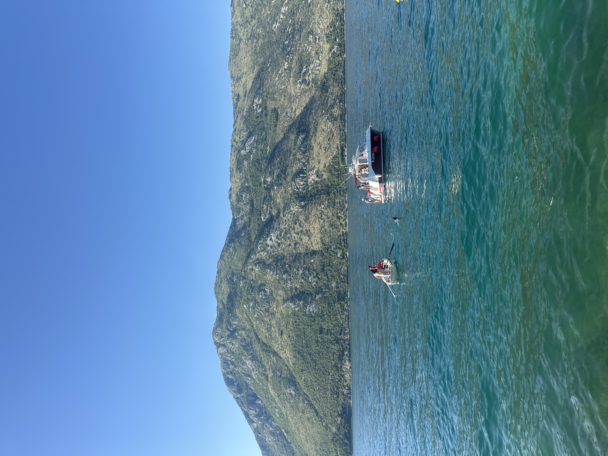 This image captures a daytime scene in the Bay of Kotor, near Prčanj, Montenegro, characterized by calm teal-blue waters and imposing mountains under a clear sky.
The upper two-thirds of the frame consists of a cloudless, vibrant blue sky. Below, a large, verdant mountain range dominates the background, stretching across the horizon. The mountains are covered with a mix of dense green foliage and exposed grey rock formations.
The foreground and midground depict a calm body of water, displaying a gradient of deep teal and blue, indicating depth and clarity. Two boats are central to the scene. On the left, a small, white rowing boat contains two individuals. One person, wearing a red garment, is seated and engaged in rowing. Another person is seated in the bow.
Further to the right and slightly behind the rowing boat, a larger white motorboat with a dark hull stripe and red fenders is positioned. At least three individuals are visible on the deck of this motorboat, some standing. One person is in the water near the stern of the motorboat, with their head visible above the surface. Fishing rods extend from the stern. The name "ADRIANA" and a partial registration number "PR" are visible on the boat's side.
Separately, in the lower-left foreground, another individual's head is visible, partially submerged in the water. A yellow buoy is partially visible in the far-right midground. The overall impression is one of leisure activities on a sunny day in a picturesque, mountainous bay.