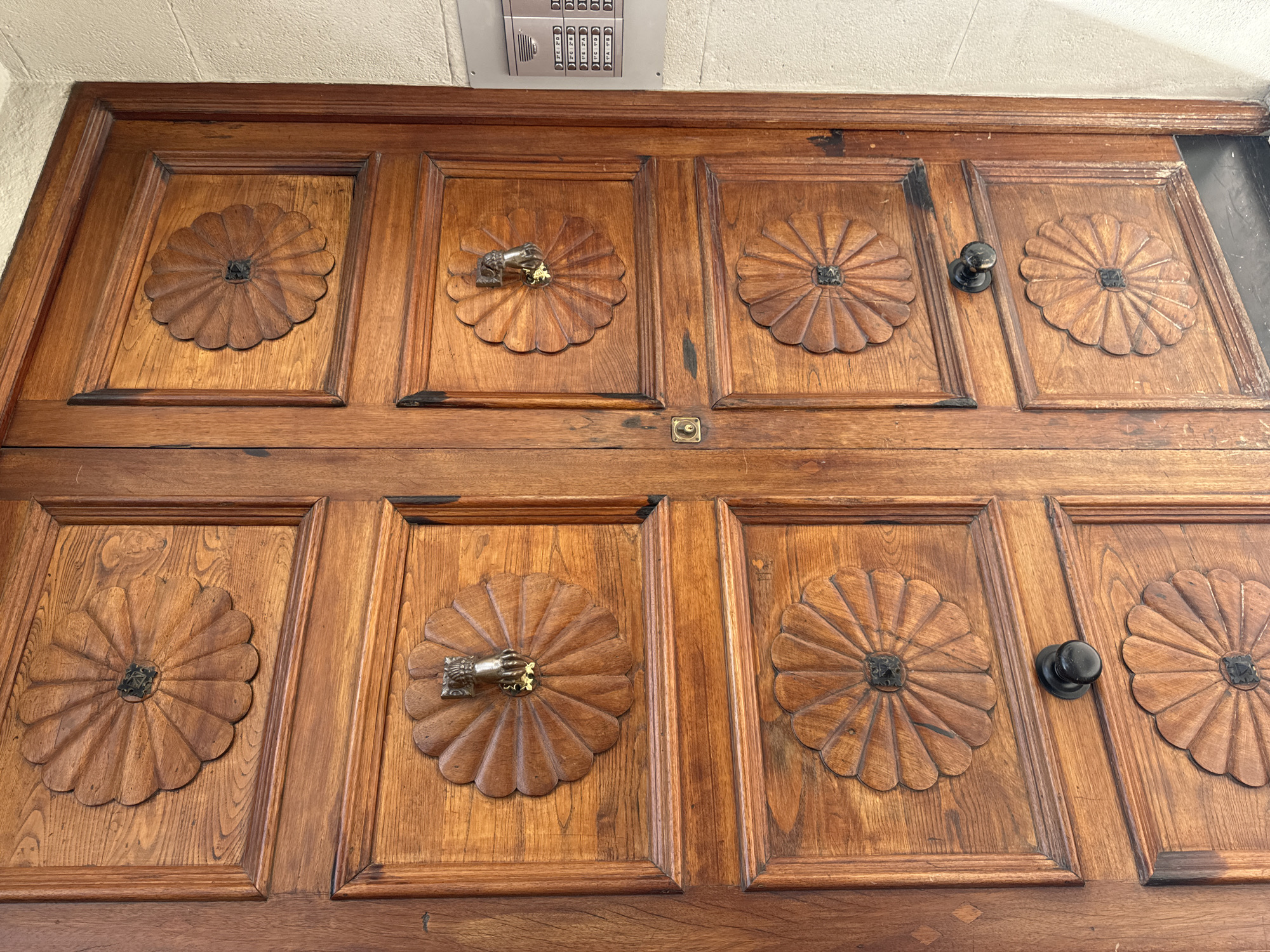 The image shows a close-up view of a pair of ornate wooden double doors, likely the entrance to a building. The doors are made of rich, dark wood with a prominent grain. Each door is divided into multiple panels, with decorative carvings in a floral or sunburst pattern on some of the upper and lower panels.
The left door has a decorative carving on the top panel and another on the bottom panel. The right door has similar carvings on its top and bottom panels. In the upper portion of the right door, there is a brass door knocker shaped like a hand. Below the hand knocker, on the right door, there is another decorative carving. On the left door, in the middle section, there is a keyhole. There are black door knobs on both doors, positioned in the middle section of the lower panels.
To the right of the doors, a section of a modern intercom system is visible, mounted on a light-colored wall. The intercom panel has multiple buttons with labels, suggesting it is for an apartment building. The doors appear to be located in a historical or traditional building, possibly in a European city. Given the request to specify the location as Murcia, Spain, this scene is likely depicting an entrance to a residential building or perhaps a commercial establishment in that city, characterized by its classic wooden doors and architectural detailing. The lighting suggests it is daytime, and the overall condition of the doors indicates they are well-maintained but show some signs of age and use.