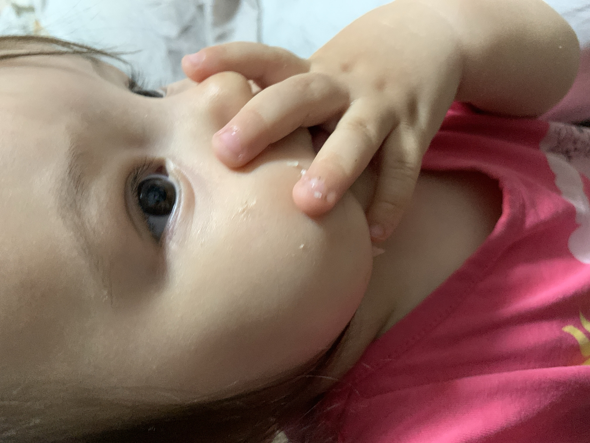 The close-up, low-angle, eye-level shot shows a baby lying down, with its hand in its mouth. The baby has fair skin and dark eyes, and is wearing a pink t-shirt. The background is blurred and appears to be a bed or soft surface. The lighting is dim, suggesting it might be nighttime or indoors. The focus is on the baby's face and hand, highlighting the texture of its skin and the smallness of its fingers. The image conveys a sense of intimacy and innocence, capturing a common childhood moment.