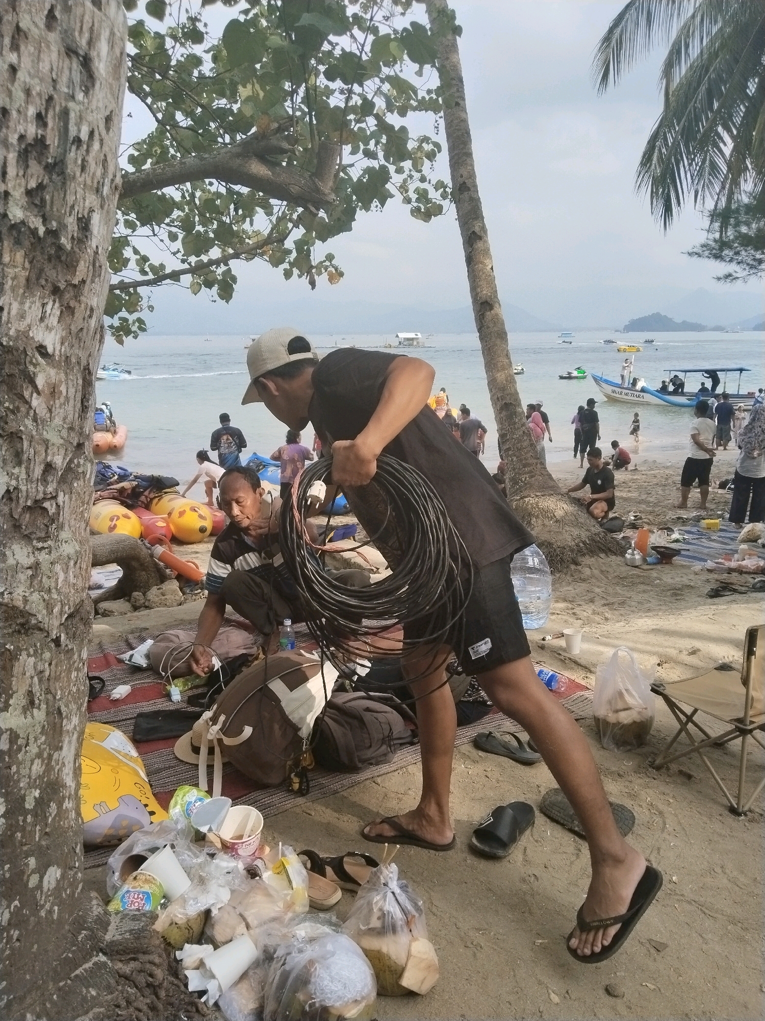 In the city of Prigi, Indonesia, a group of people are on a beach near the ocean. A man wearing a black t-shirt and shorts is standing, holding a coil of black cable. Another man, seated on a striped mat, is wearing a striped shirt and dark pants, appearing to be working with some items on the mat. Numerous inflatable buoys, some yellow and some orange, are scattered on the sand. In the background, several boats are visible on the water, with people boarding and disembarking. The beach is sandy, with some vegetation and palm trees present. A large tree trunk is prominent in the foreground on the left side of the image. Various bags, containers, and personal items are strewn across the sand, suggesting a gathering or picnic. The sky is overcast.