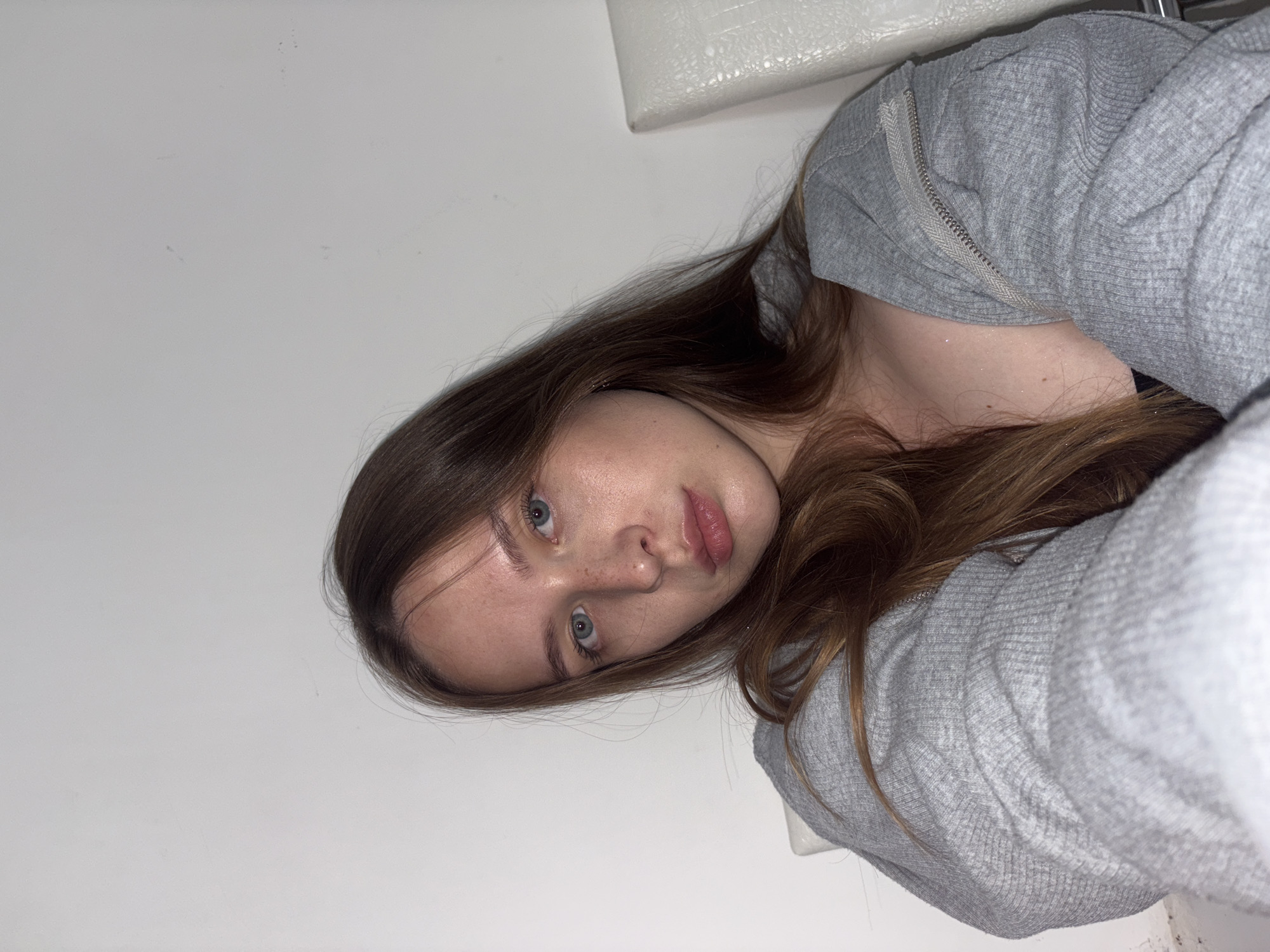 A young woman with long brown hair and blue eyes is looking directly at the camera. She is wearing a grey zip-up hoodie. She has freckles on her face. She is sitting in what appears to be an indoor setting, with a white wall behind her and a glimpse of a white chair to her right. There is no indication of the city of Chaikovsky, Russia in the image.