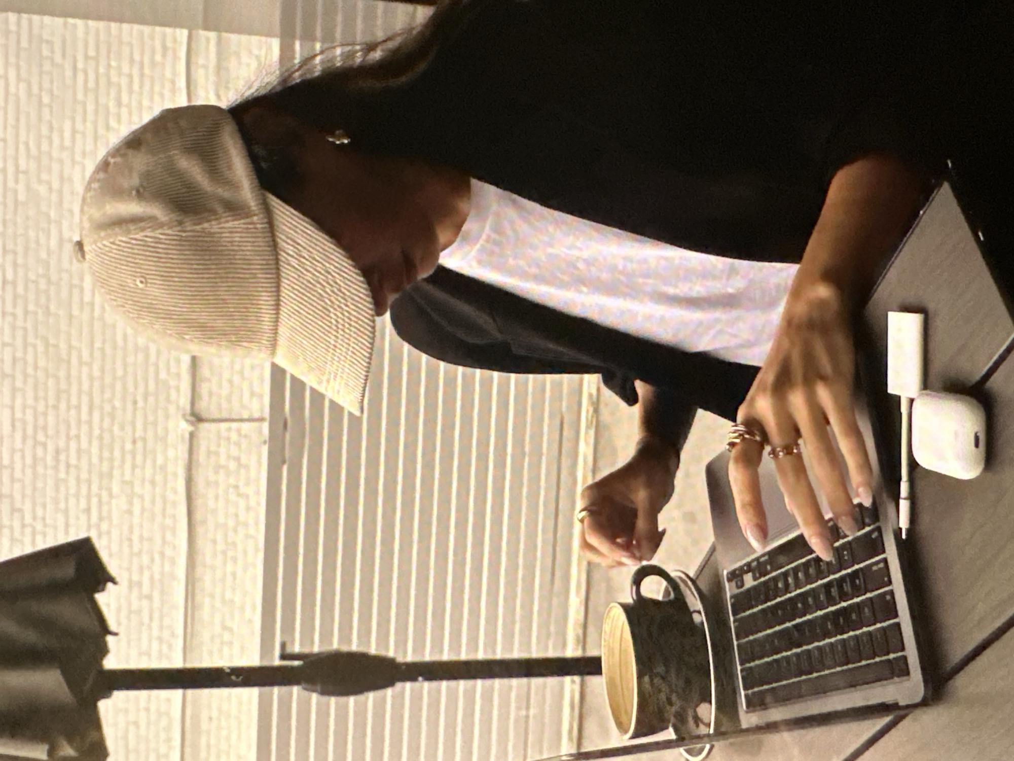 A person wearing a light-colored baseball cap, a white t-shirt, and a black jacket is sitting at a table, typing on a laptop. Their hands are adorned with multiple rings. A coffee mug is on the table to the left of the laptop, and a white dongle with an audio cable is plugged into the laptop's right side. A pair of white earbuds is lying near the laptop. The location is outdoors, with a vertically ribbed surface behind the person, and a partial view of an umbrella stand on the left. The city of Yerevan, Armenia, is not identifiable in the image.