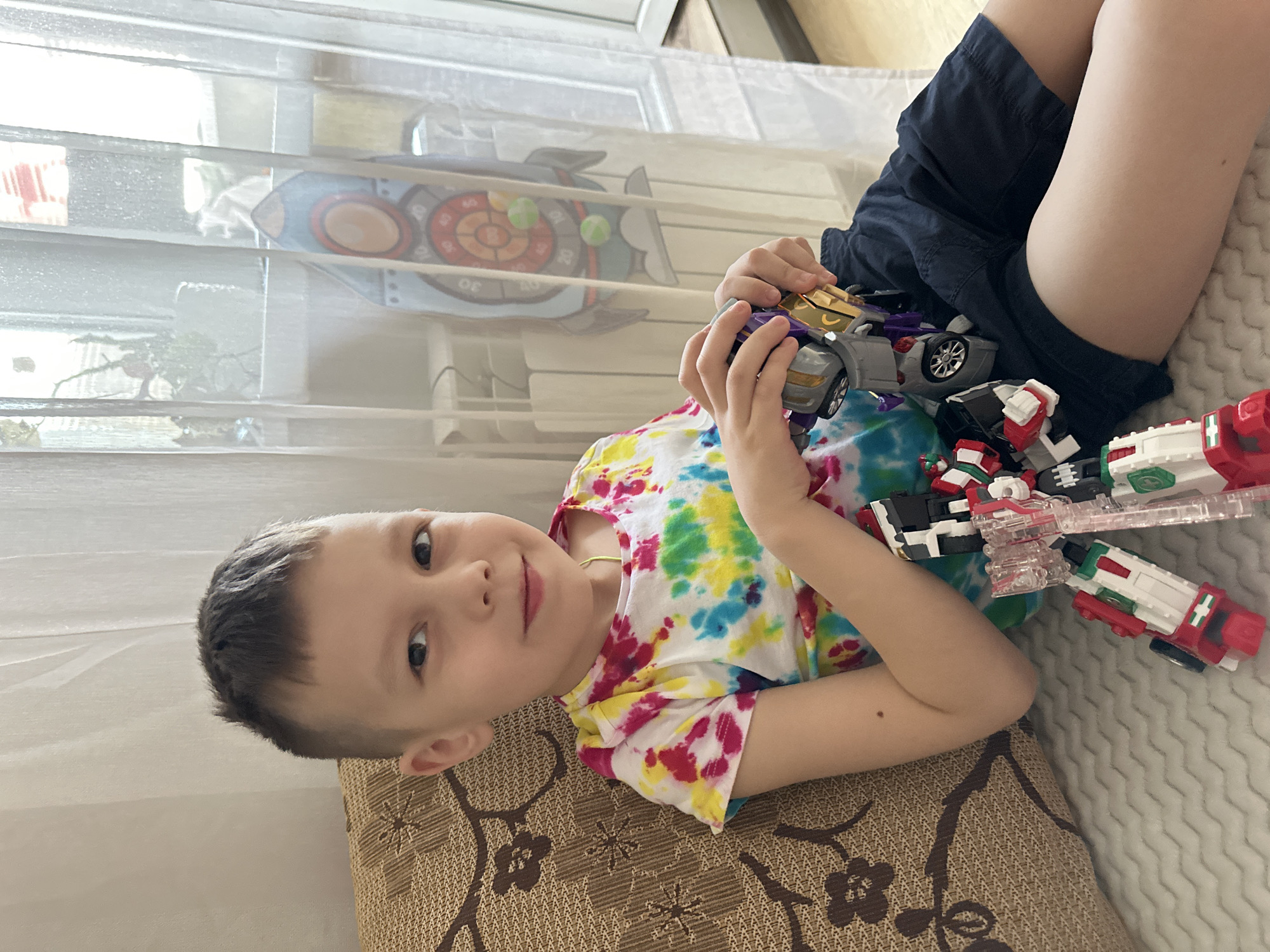A young boy, appearing to be around 6-8 years old, is lying on a brown and tan patterned pillow, seemingly on a bed or couch. He is wearing a tie-dye shirt with colors of white, yellow, green, blue, and pink. He is holding several toy vehicles, including a purple and grey car, and a set of red and white vehicles. He is looking directly at the camera and smiling slightly. The background includes a window with sheer curtains. Behind the curtains, an object resembling a dartboard is visible. The location is speculated to be Tomilino, Russia based on the user request. The boy appears to be relaxed and engaged in playing with his toys. He is wearing dark blue shorts.