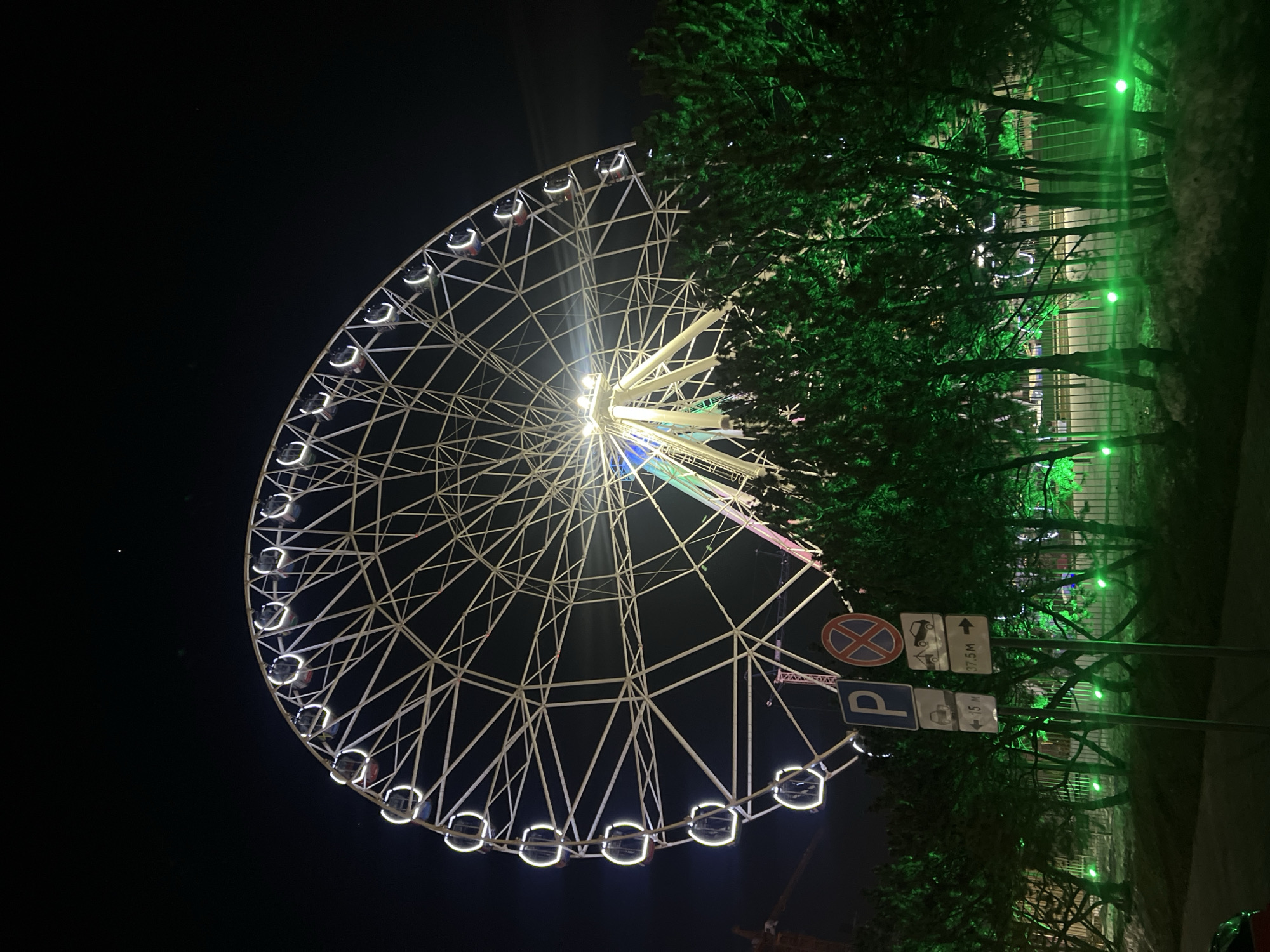 The image depicts a Ferris wheel at night in Kazan, Russia. The wheel is illuminated with white lights outlining its structure and passenger cabins. Its center is lit with multiple colors. To the right of the Ferris wheel, trees are illuminated with green lights. Street signs for parking and towing zones are visible near the trees. The sky is dark, indicating a nighttime setting. A construction crane can be seen in the background under the Ferris wheel.
