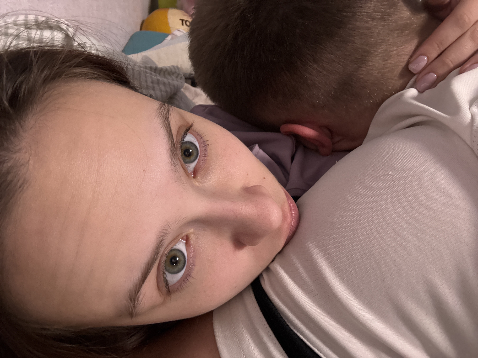 An intimate, close-up indoor portrait features a woman and a child in what appears to be a bedroom setting in Прикамский, Russia. The woman, positioned on the left side of the frame, lies on her side, looking directly into the camera. She has striking green eyes, subtle makeup, and dark hair pulled back, with wisps visible around her forehead. Her lips are slightly parted. She is wearing a white top with a black stripe, and a portion of her shoulder and chest is visible.
Resting against the woman's side is a child, whose head is visible from the back, indicating short, light brown hair. The child's ear is peeking out from behind his head. The woman's hand, adorned with light pink manicured nails, gently cups the back of the child's head, suggesting a comforting embrace. The child appears to be wearing purple clothing.
The background is softly blurred but reveals bedding, including a striped grey and white blanket or sheet, and a yellow and white object, possibly a pillow or toy, on which the letters "TO" are partially visible. The lighting is soft and subdued, typical of an indoor evening or early morning scene, creating a peaceful and tender atmosphere.