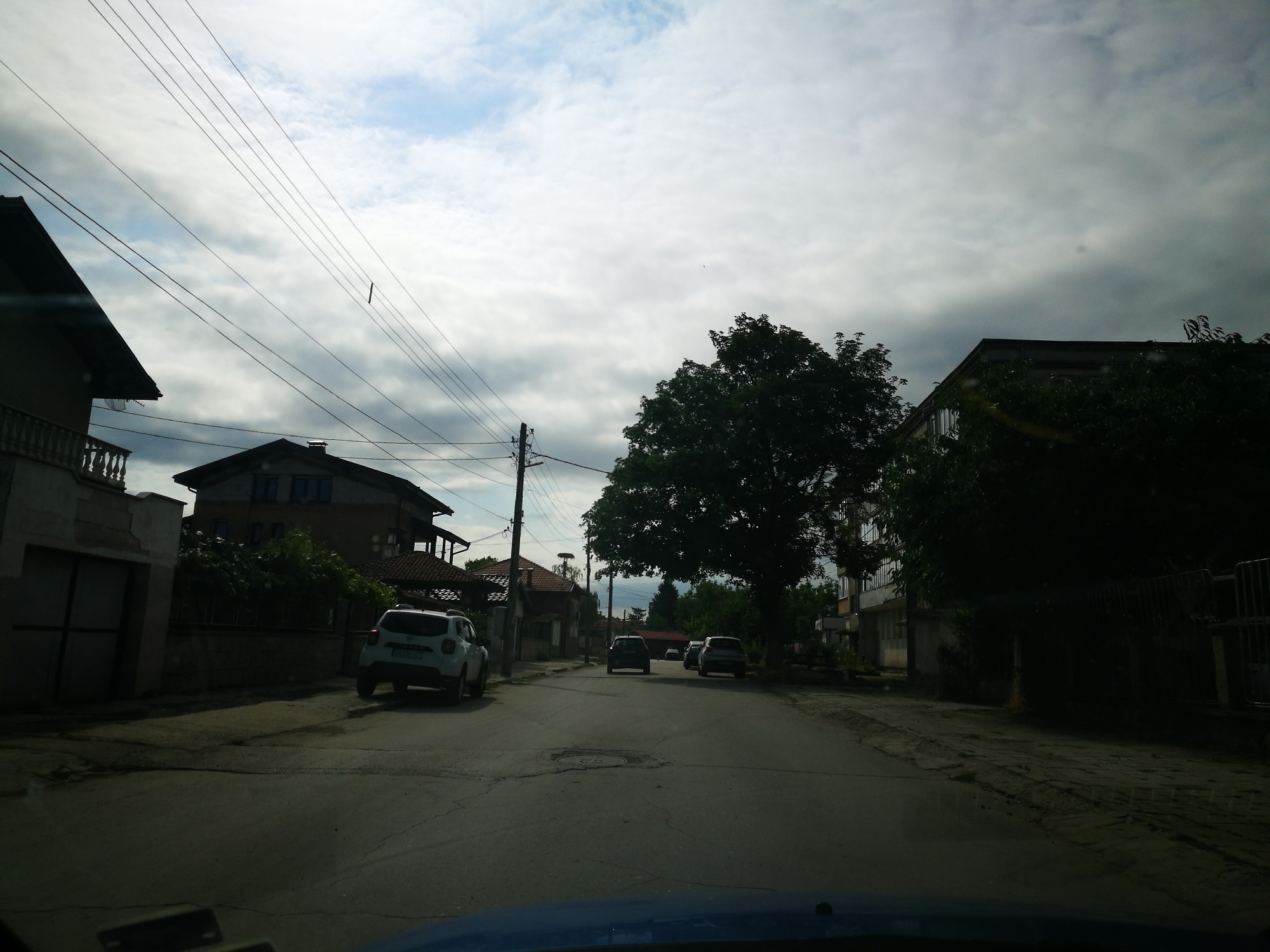 The image shows a street scene in Ihtiman, Bulgaria. The perspective is from inside a vehicle looking forward. The street is paved and appears to be in a residential area with houses on either side. There are several cars on the road, including a white Dacia Duster parked on the left and two cars further down the street. A large tree is prominent in the middle of the frame. Overhead power lines crisscross the sky, which is partly cloudy. The architecture of the houses suggests a traditional Eastern European style, with tiled roofs and balconies. In the background, there is a stork's nest on a pole. The lighting suggests it is daytime, possibly late afternoon or early morning, given the shadows.